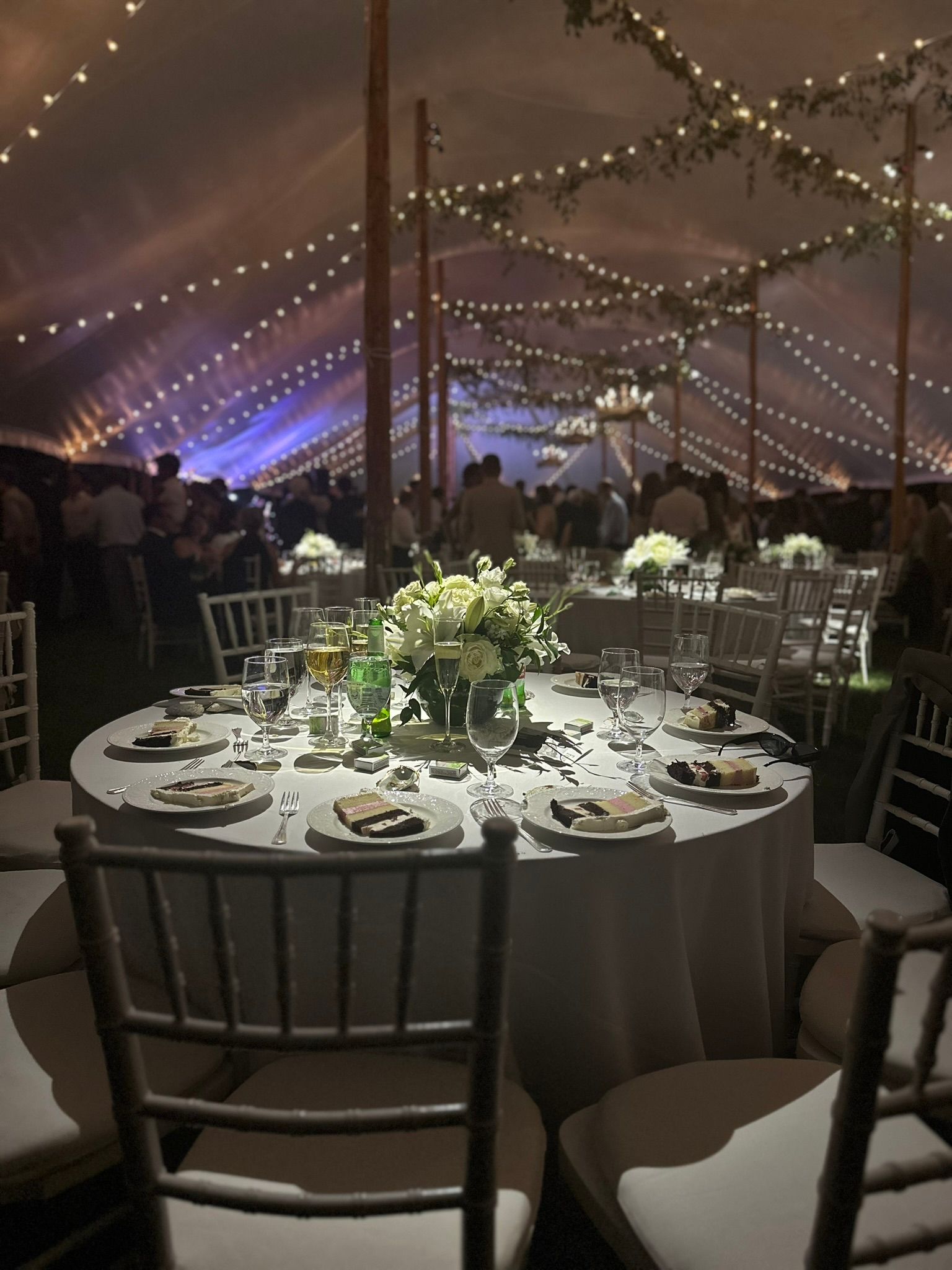 Round table set for a formal event under a tent with string lights and floral centerpiece.