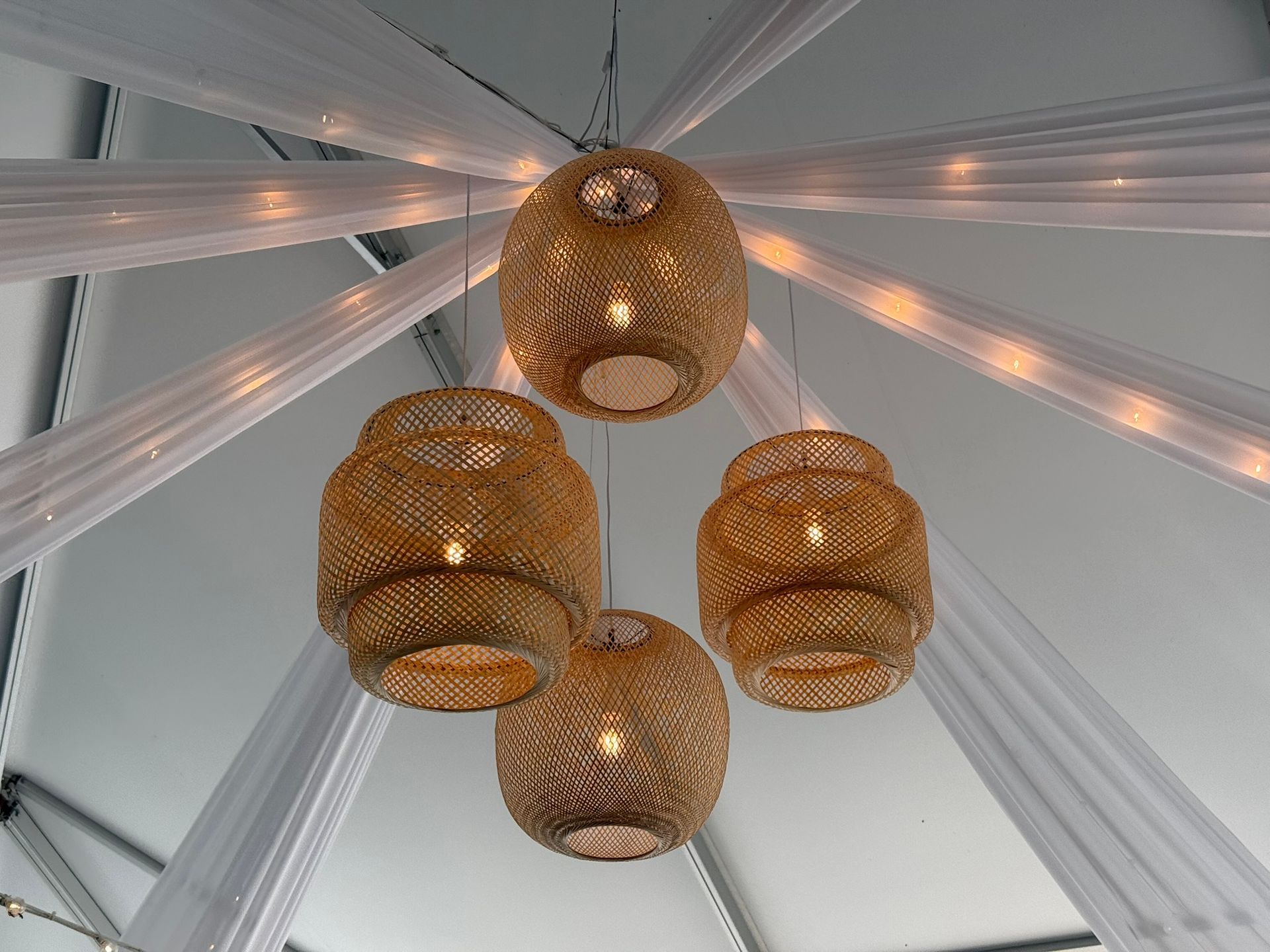 Four woven gold pendant lights hanging from a white ceiling with draped fabric and small lights.