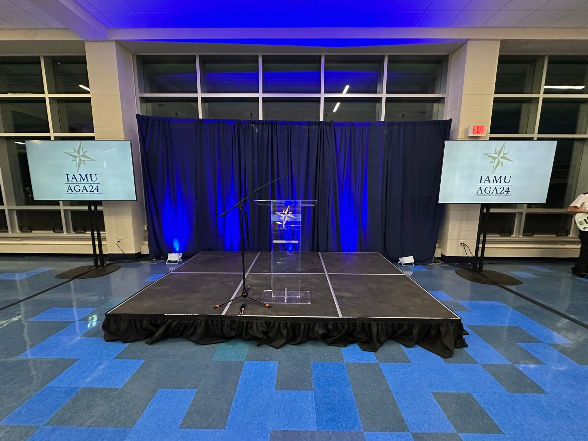 Stage with podium, blue backdrop, screens displaying text. Blue and white floor.