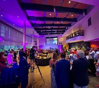 Event hall with people, food tables, stage, and camera under purple lighting.
