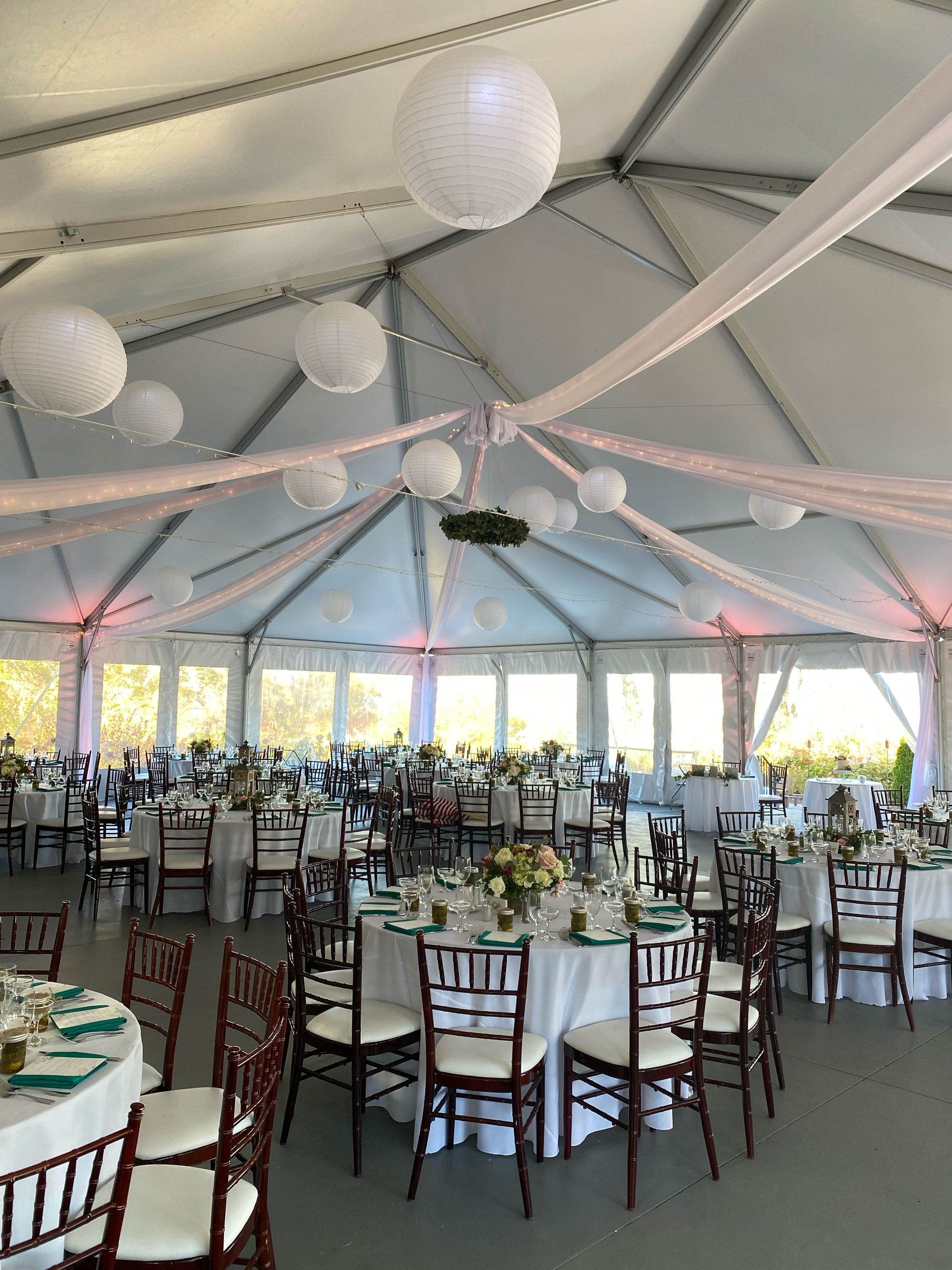 Wedding reception in a tent with round tables, chairs, and decorations. White lanterns hang from the ceiling.