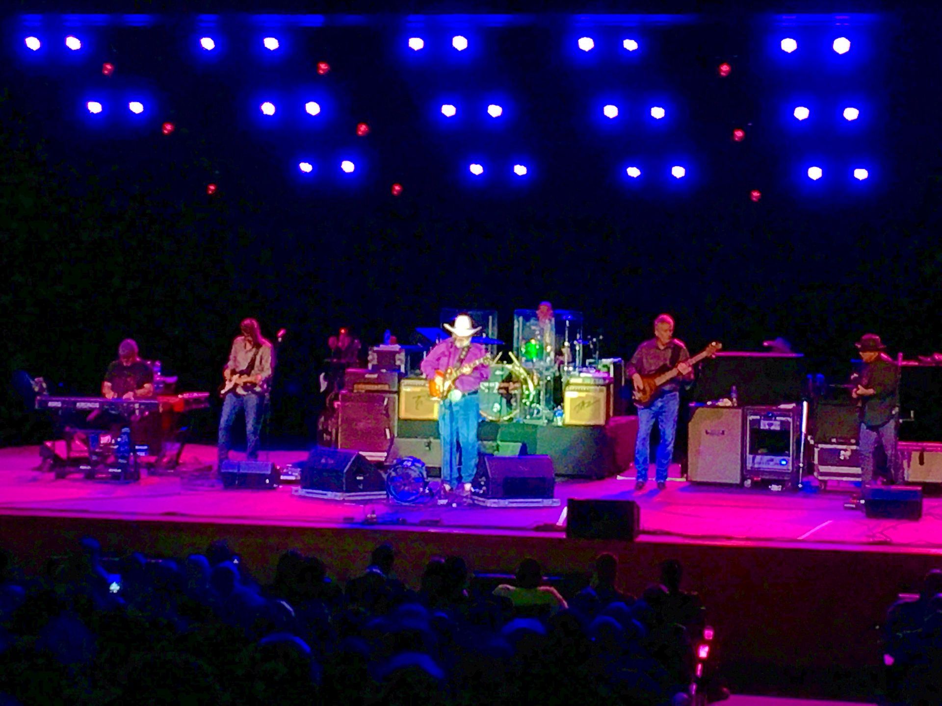 Band performing on stage under blue and red lights. Musicians play instruments, audience in front.