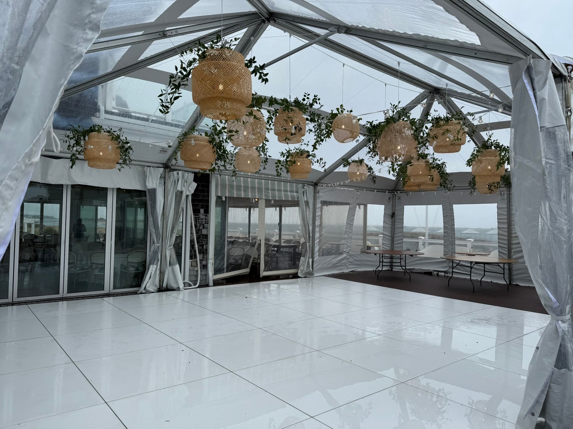 Inside clear-roofed event tent, white dance floor, decorated with hanging woven lights, greenery, and glass walls.