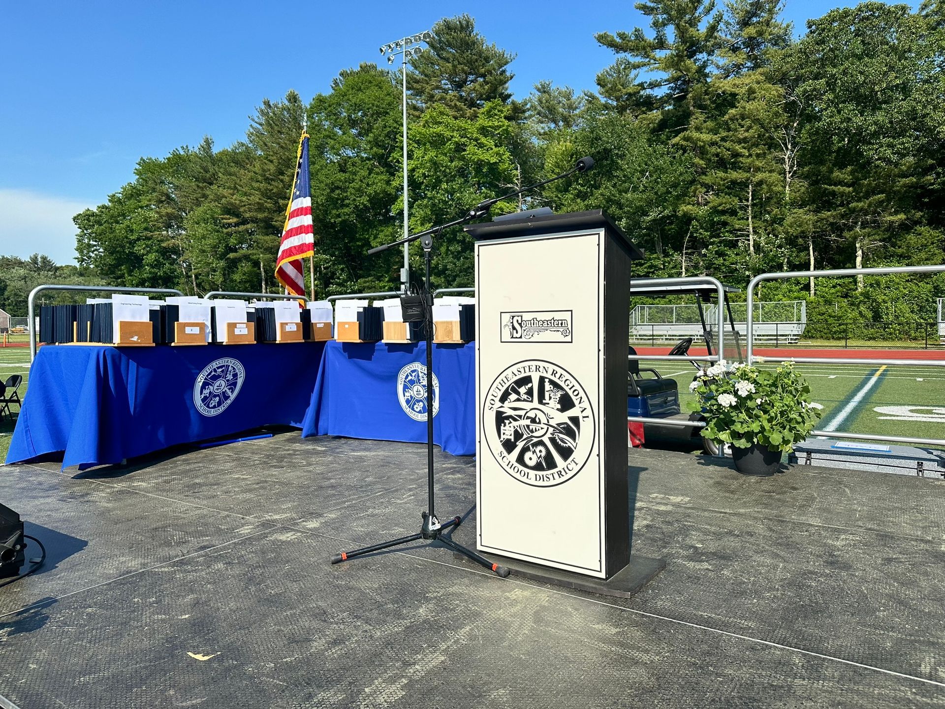 Podium with microphones in front of a table with boxes and US flag. Football field and trees in the background.