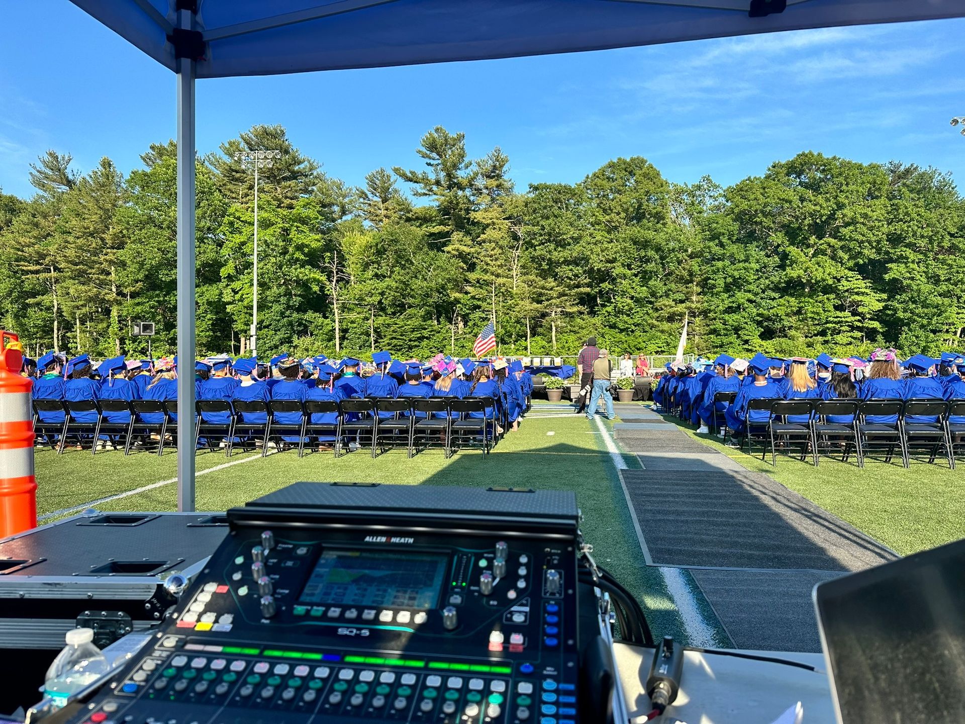 A graduation ceremony set up on a grassy field. Students in blue gowns sit in rows, facing a speaker's podium. Sound equipment in foreground.