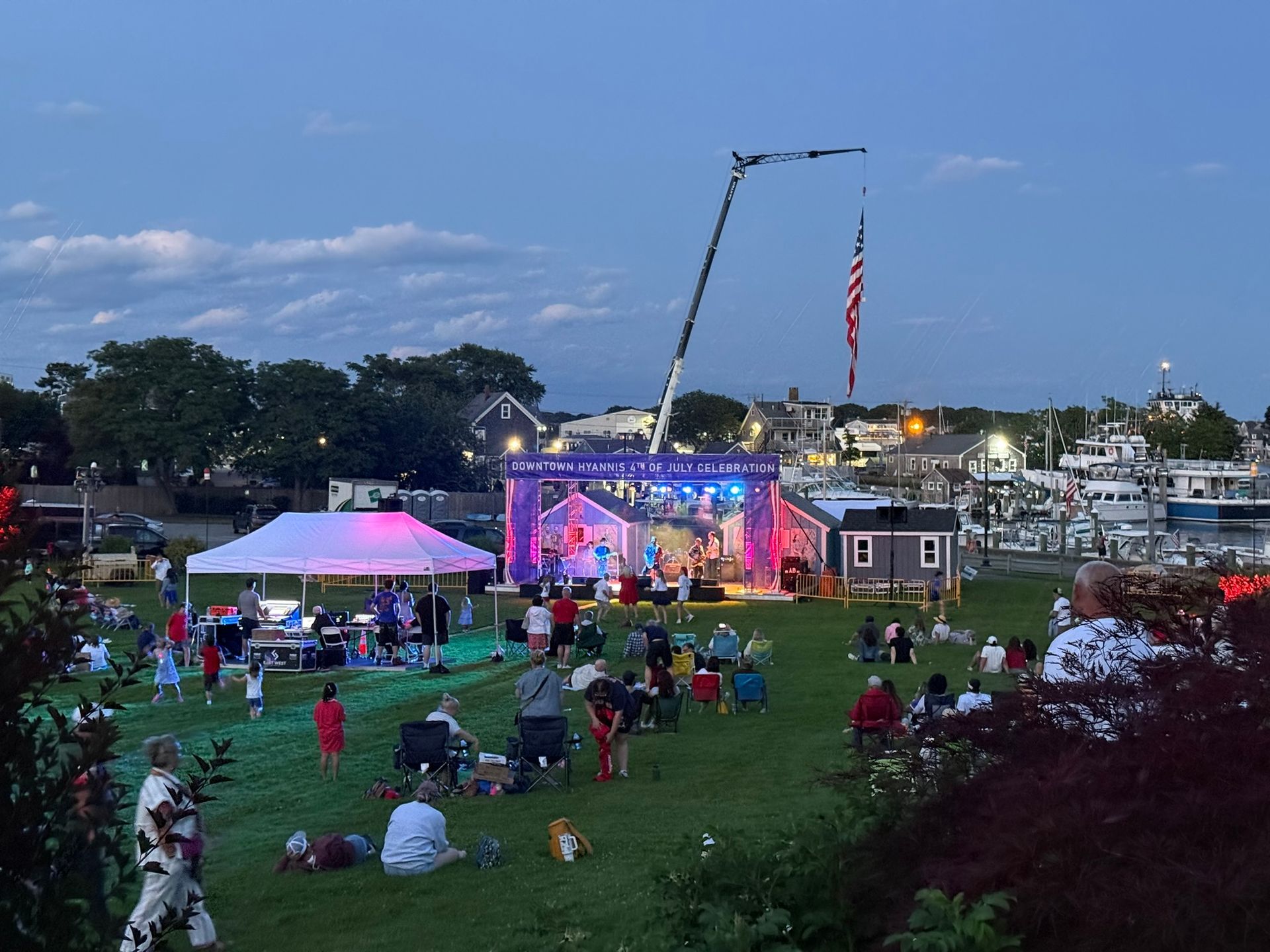 Concert on a grassy waterfront, stage lit with colorful lights, audience seated. American flag on tall crane.