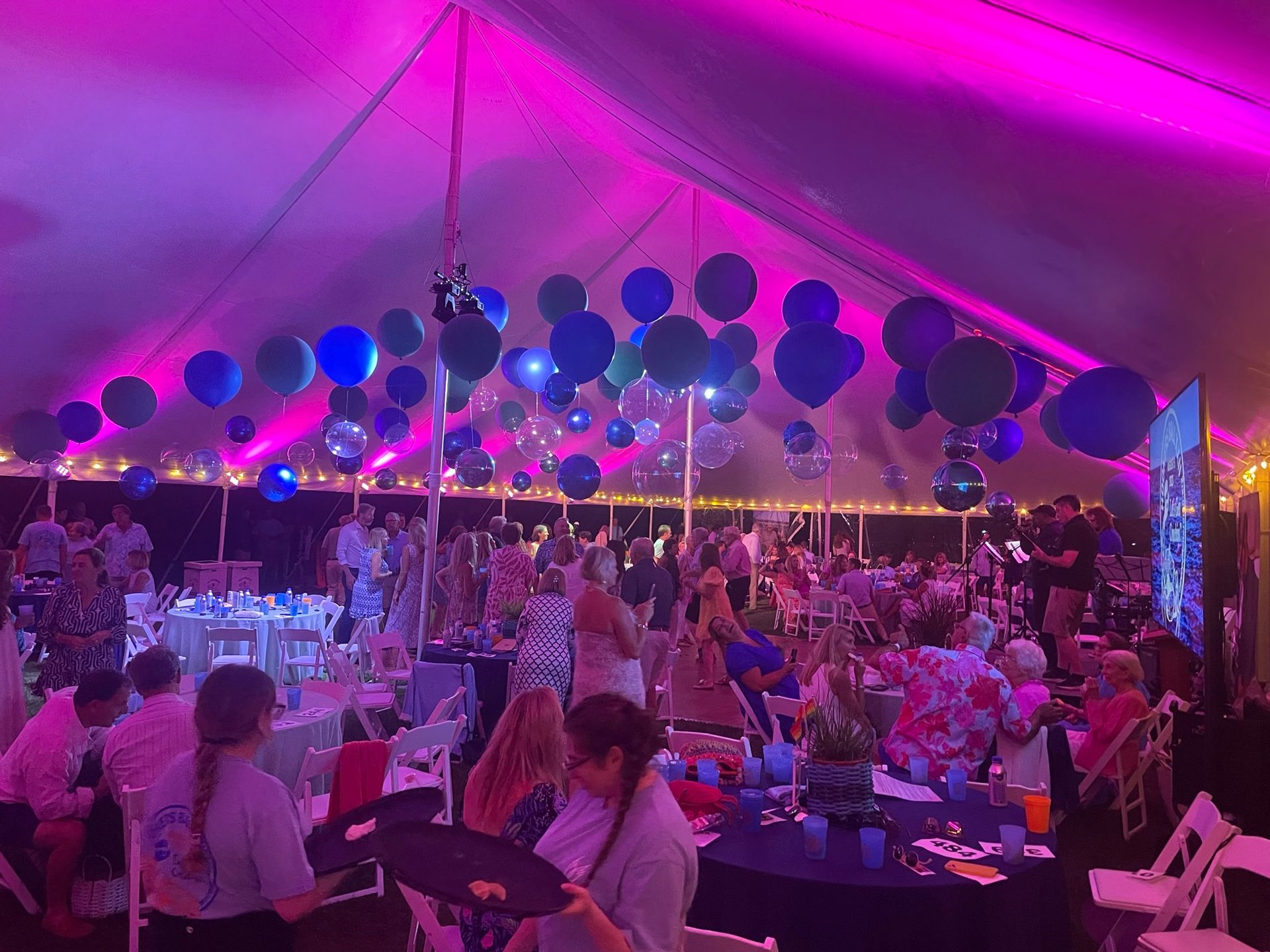 Party tent interior with guests, tables, balloons, and pink and blue lighting.