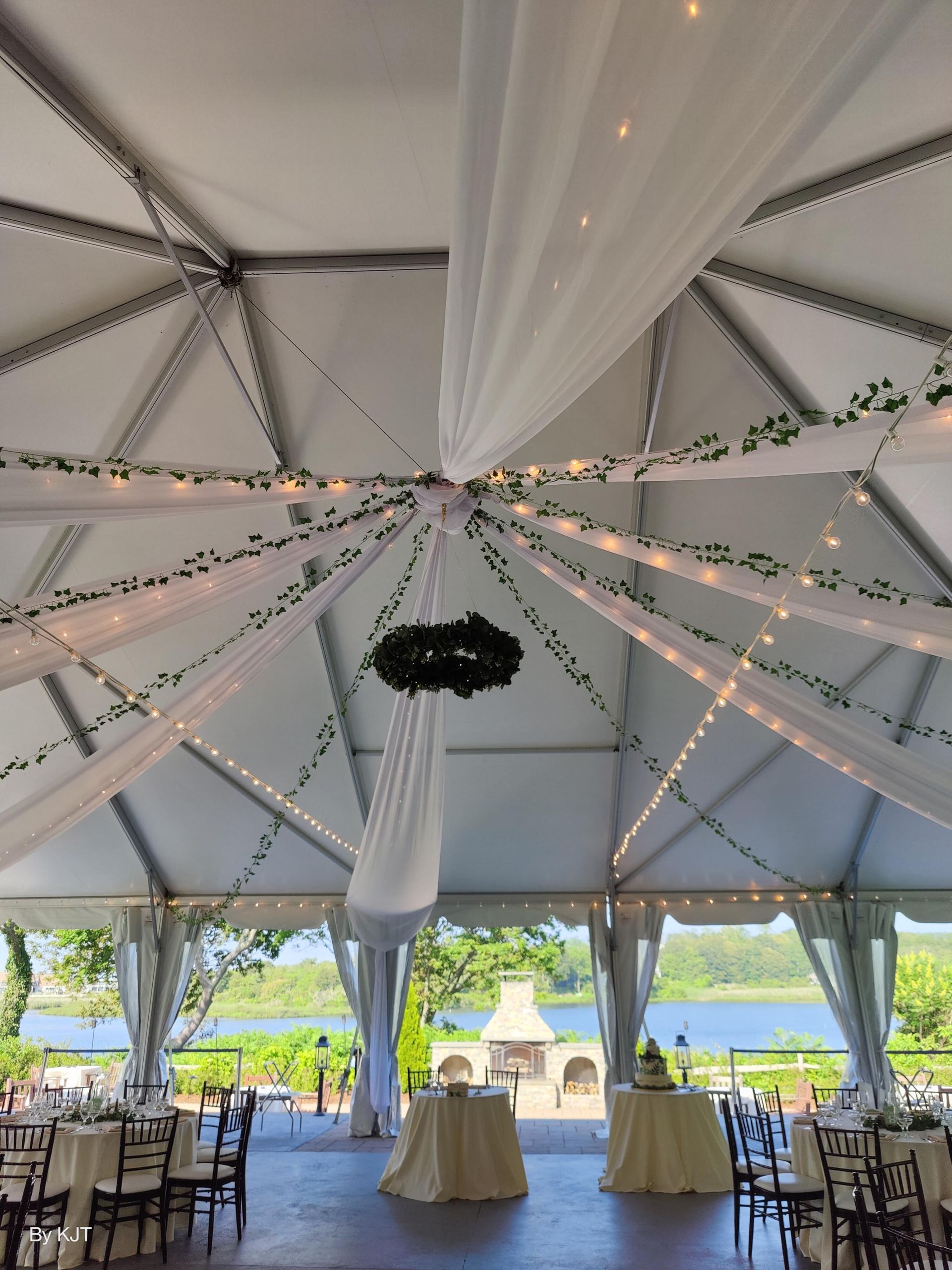 Wedding reception tent, decorated with white fabric, fairy lights, and greenery, overlooking a lake.