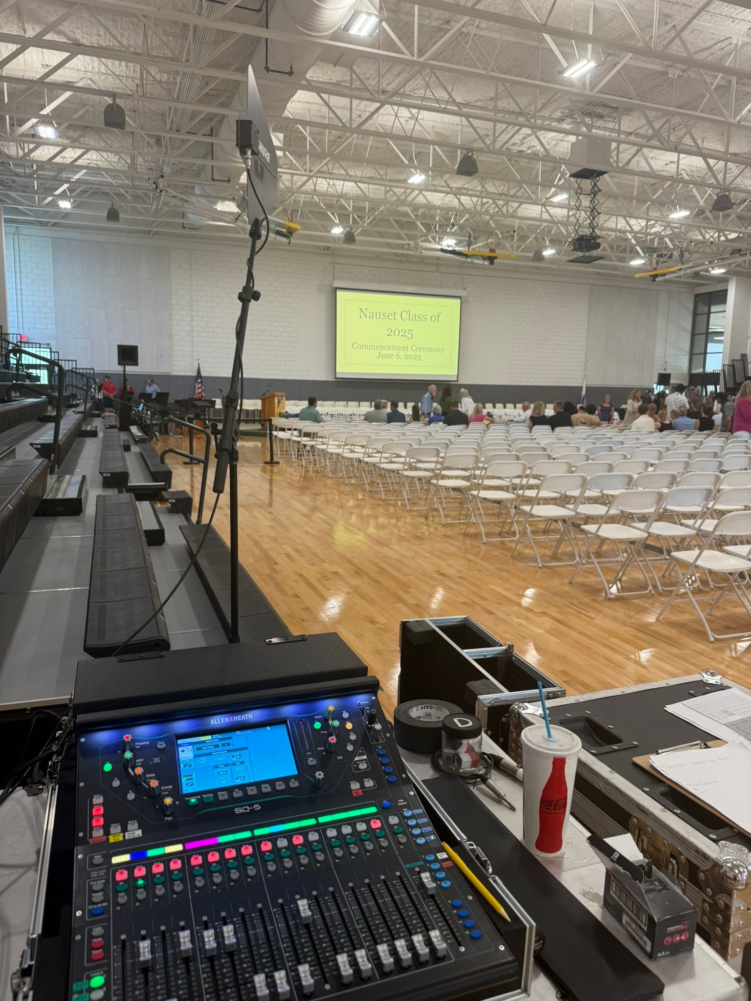 Soundboard set up in a gymnasium for a presentation. Rows of white chairs face a screen with text, people seated in the distance.
