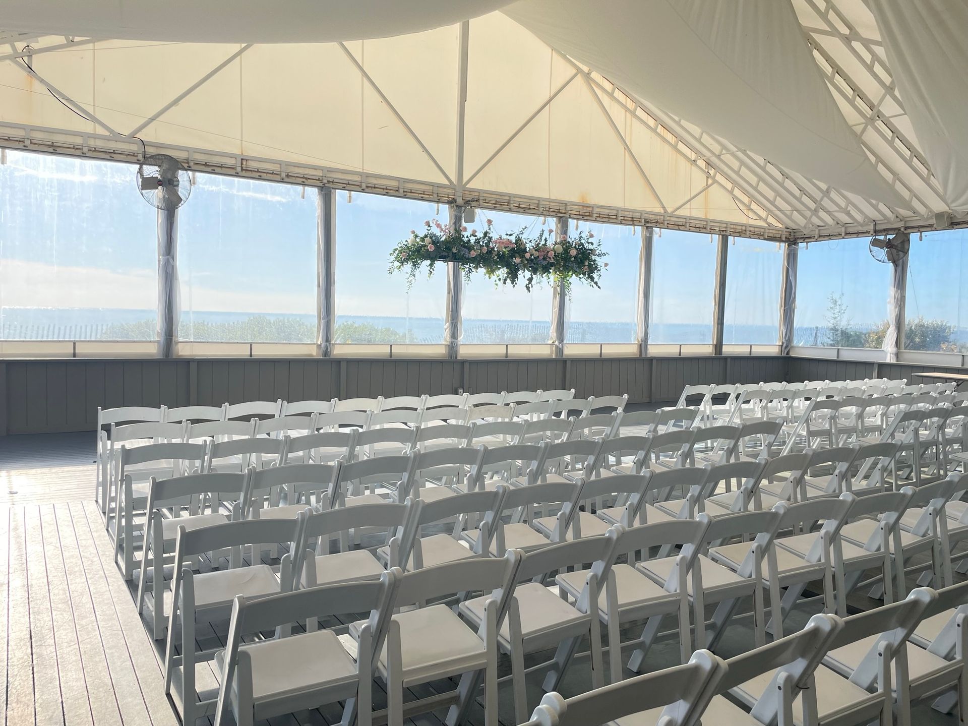 Rows of white chairs arranged inside a tent with a floral decoration, overlooking a landscape.