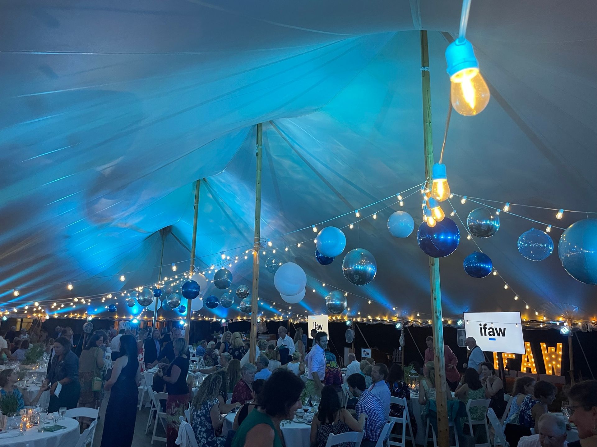 Large event tent with guests. Blue lighting, string lights, and balloons decorate the space.