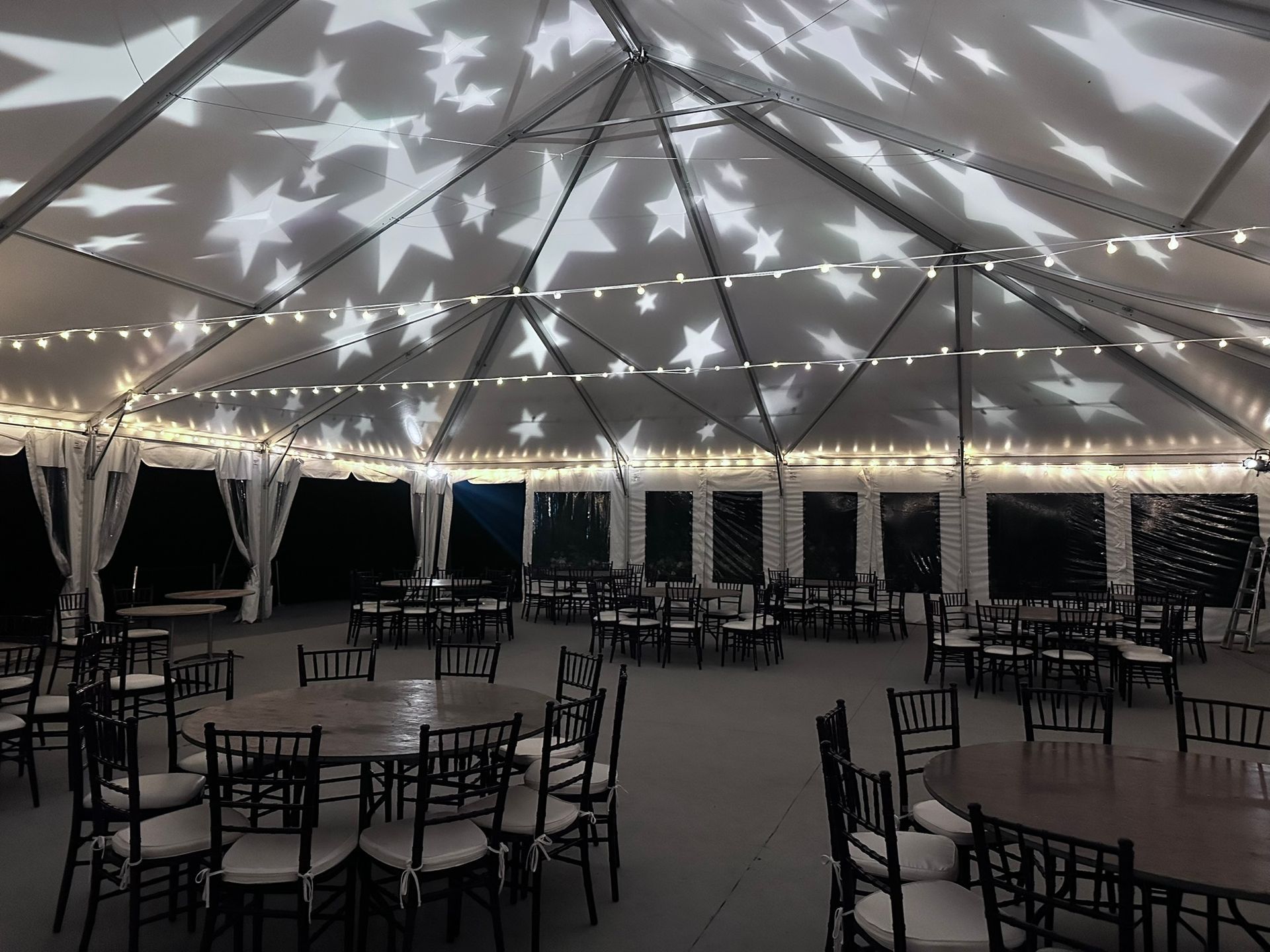 Star-lit tent interior set for an event; round tables with black chairs; string lights and draped curtains.