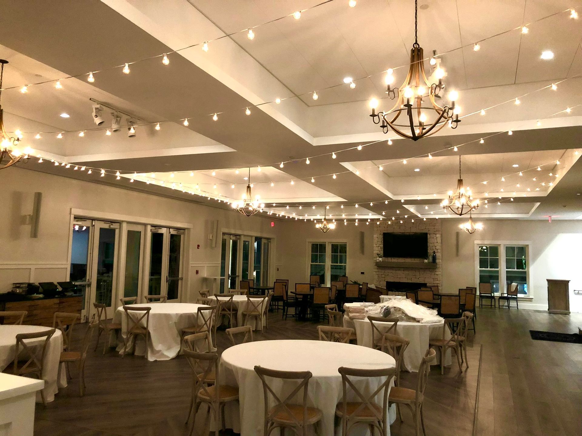 An event space with round tables, chairs, chandeliers, string lights, and a fireplace.
