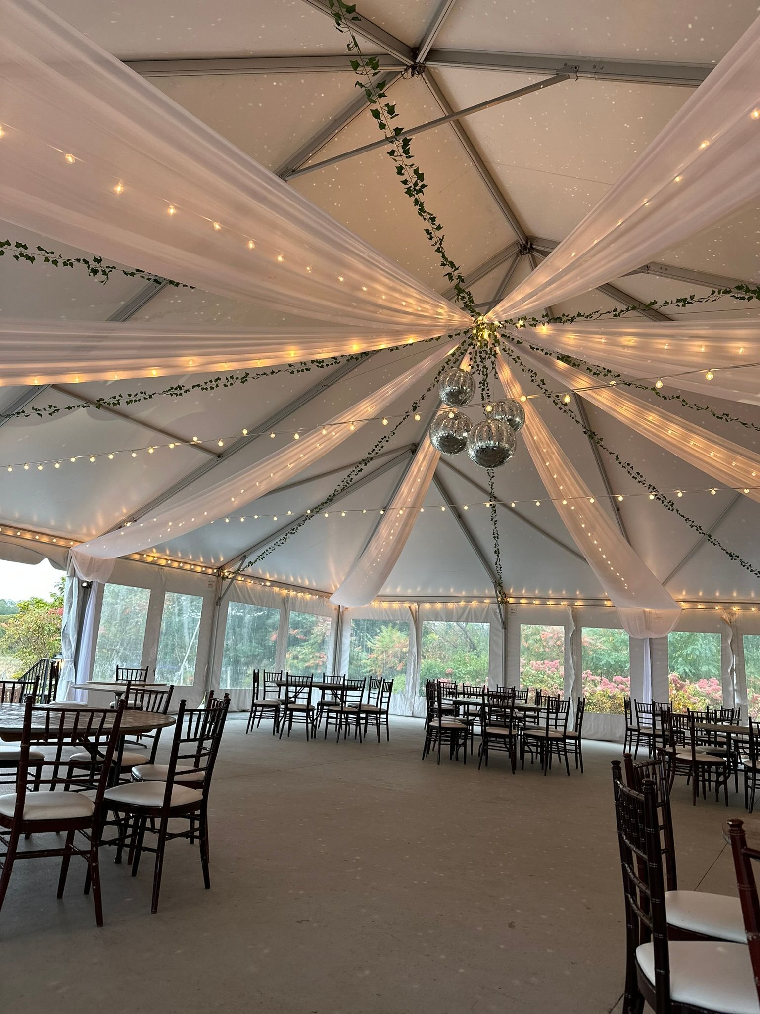 Wedding reception tent with tables, chairs, sheer fabric, and string lights.