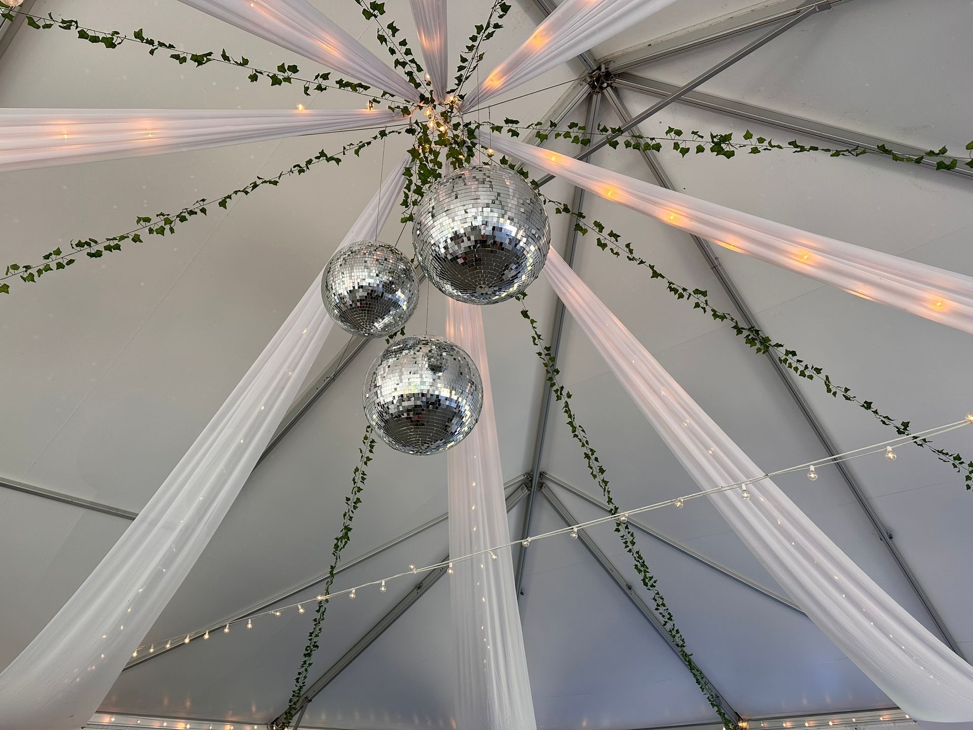 Three disco balls hanging from a white tent ceiling with draped fabric and string lights.