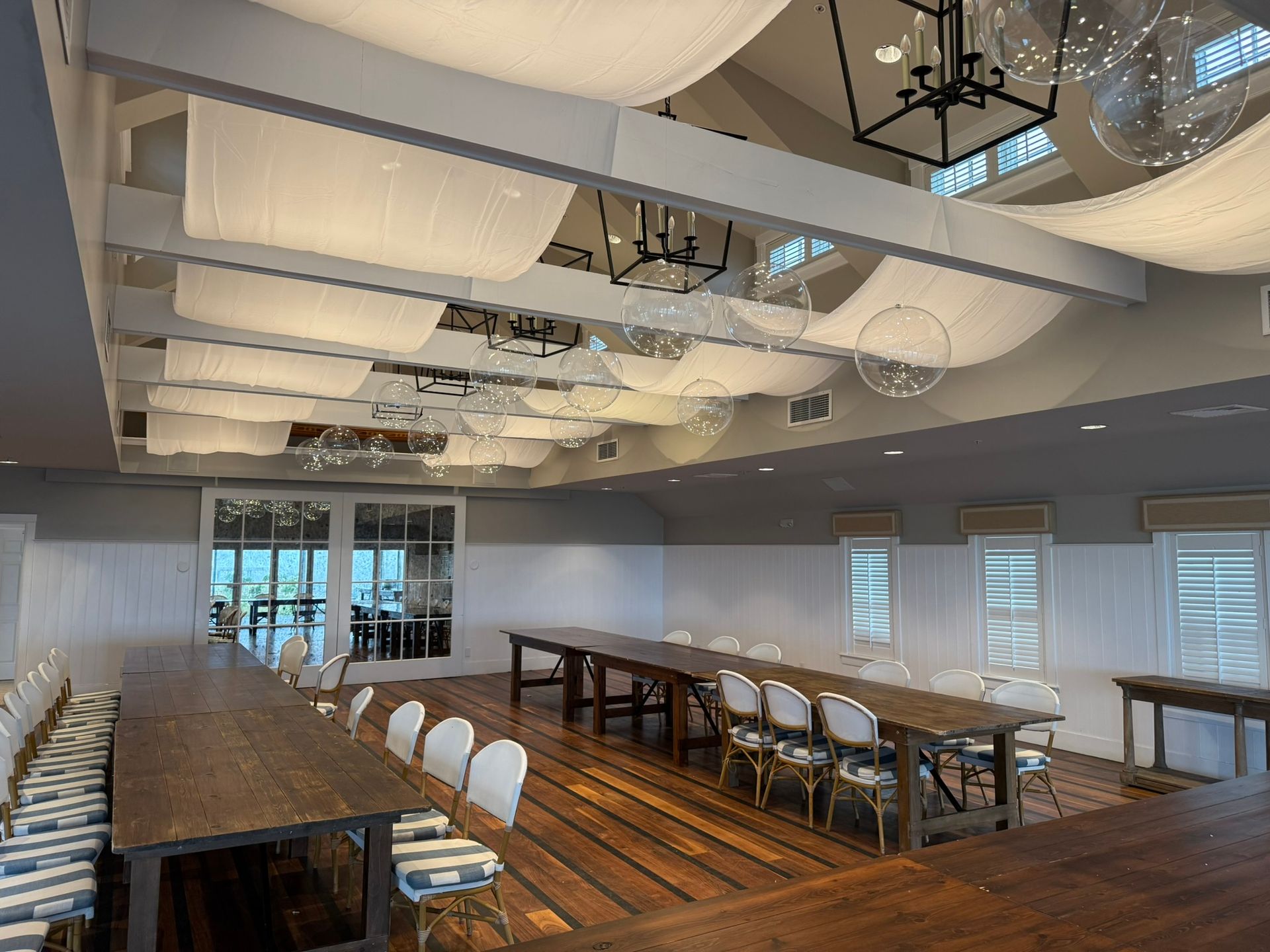 Long wooden tables with white chairs in a brightly lit event space with draped fabric ceiling.
