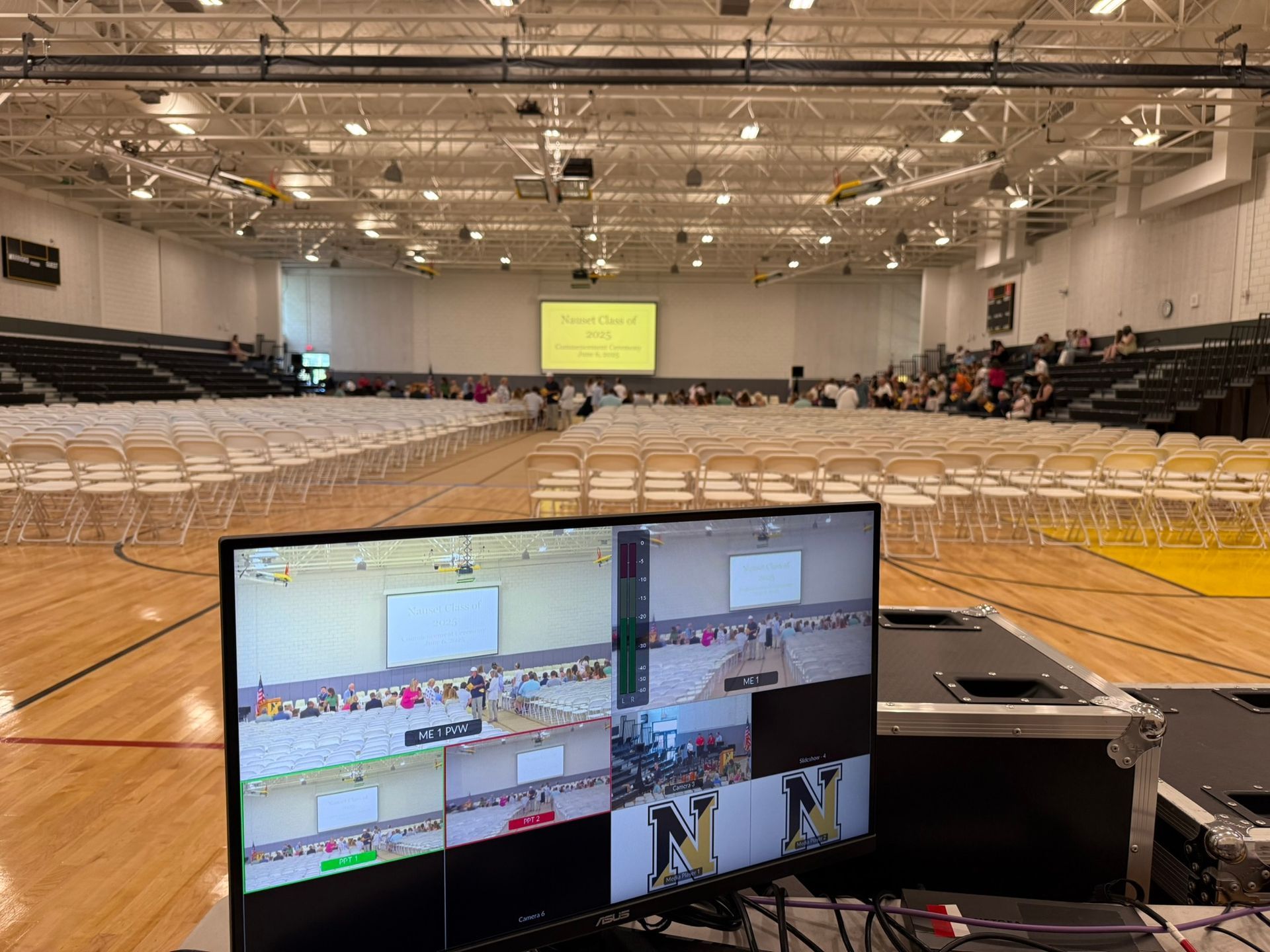 A gymnasium set up for an event, with chairs and a screen. A control monitor is in the foreground.