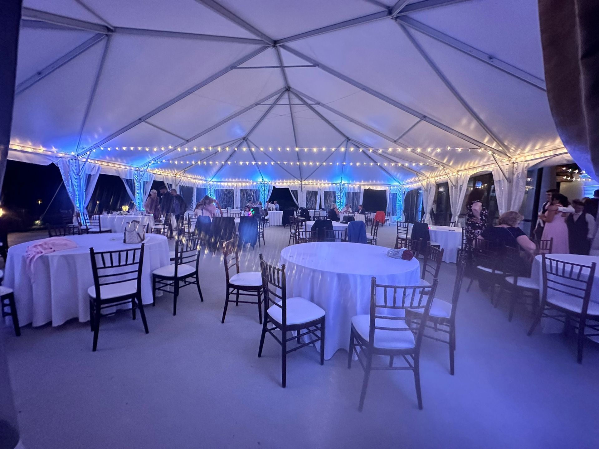 Outdoor event tent with tables, chairs, and string lights. People are seated and socializing.