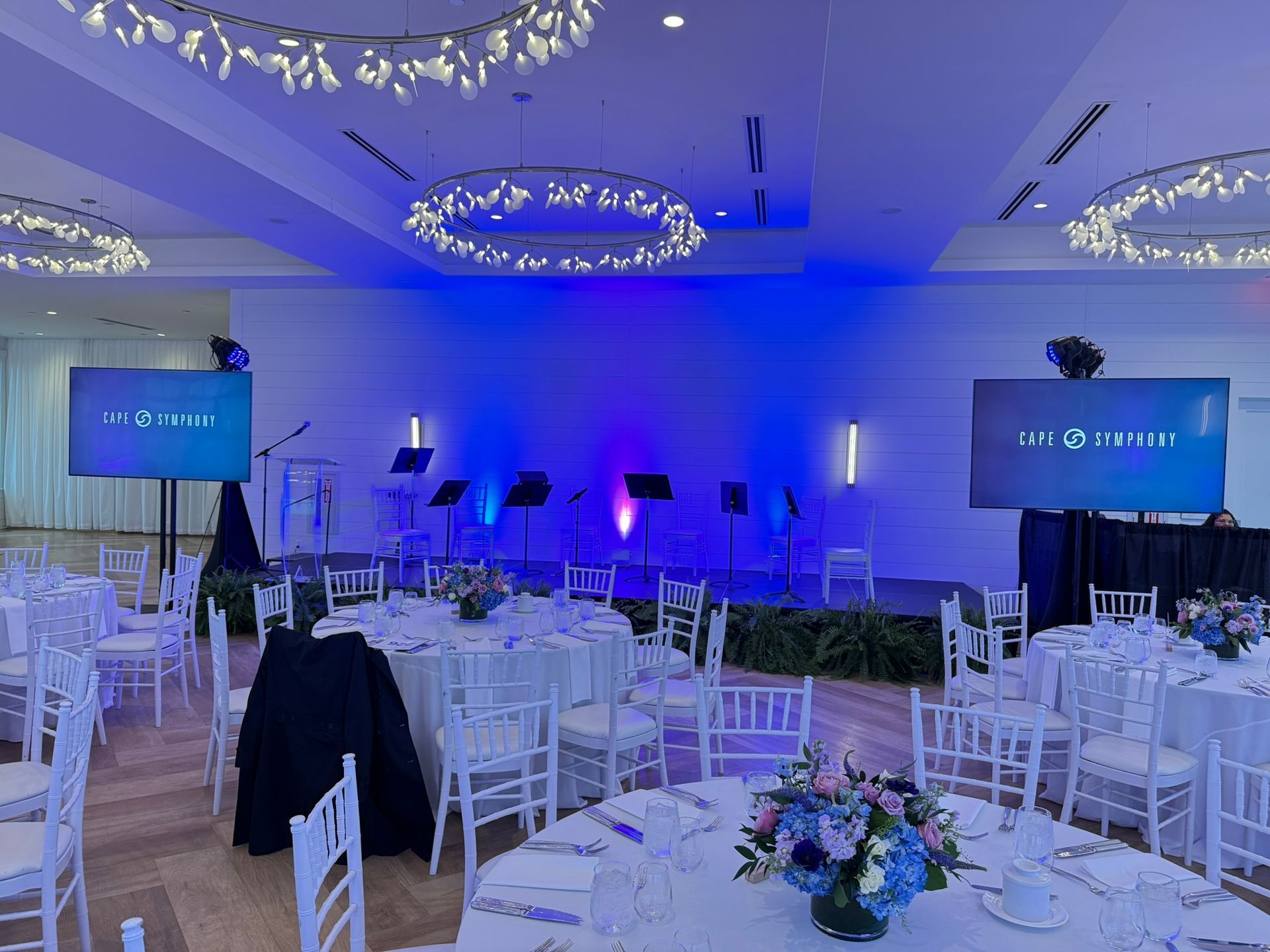 Banquet hall set for an event with round tables, stage, screens, and blue lighting.