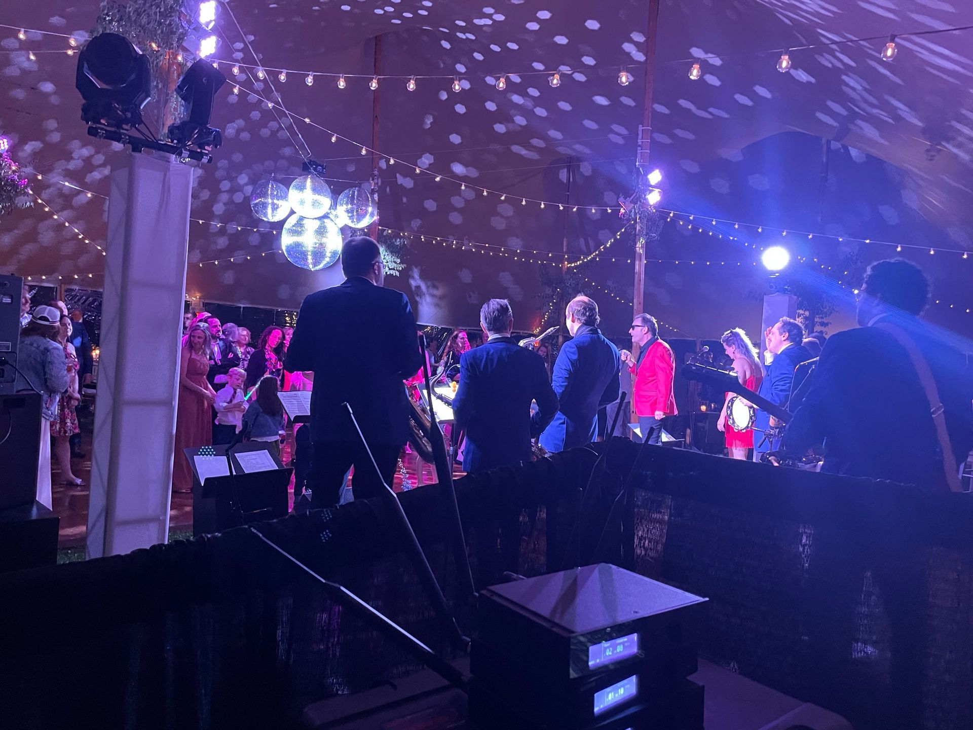 Band playing on stage at a party under a tent with disco balls and lights. People are dancing and tables with guests are visible.