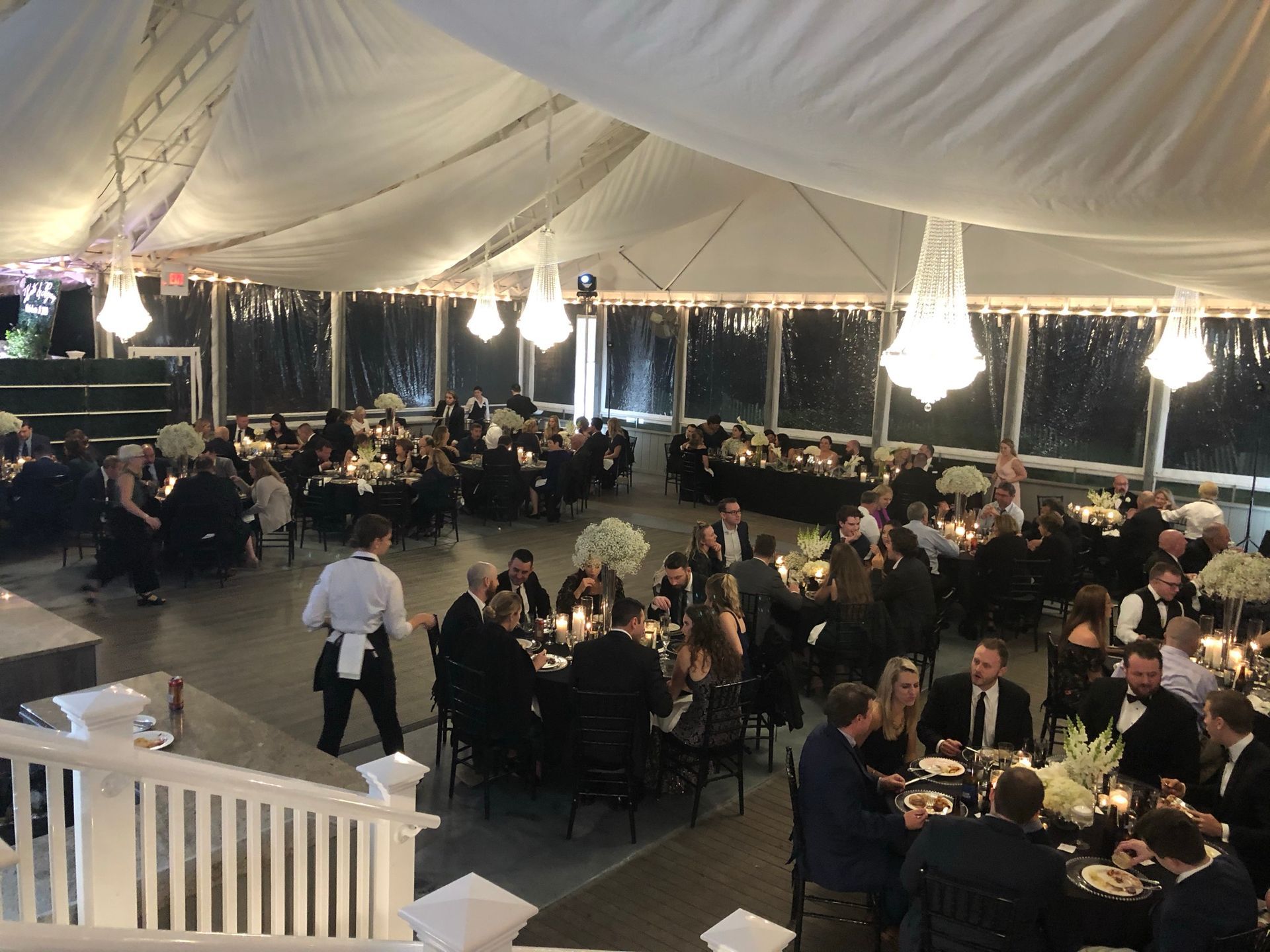 Inside a tent: guests seated at black tables, chandeliers hang above, server walks by.