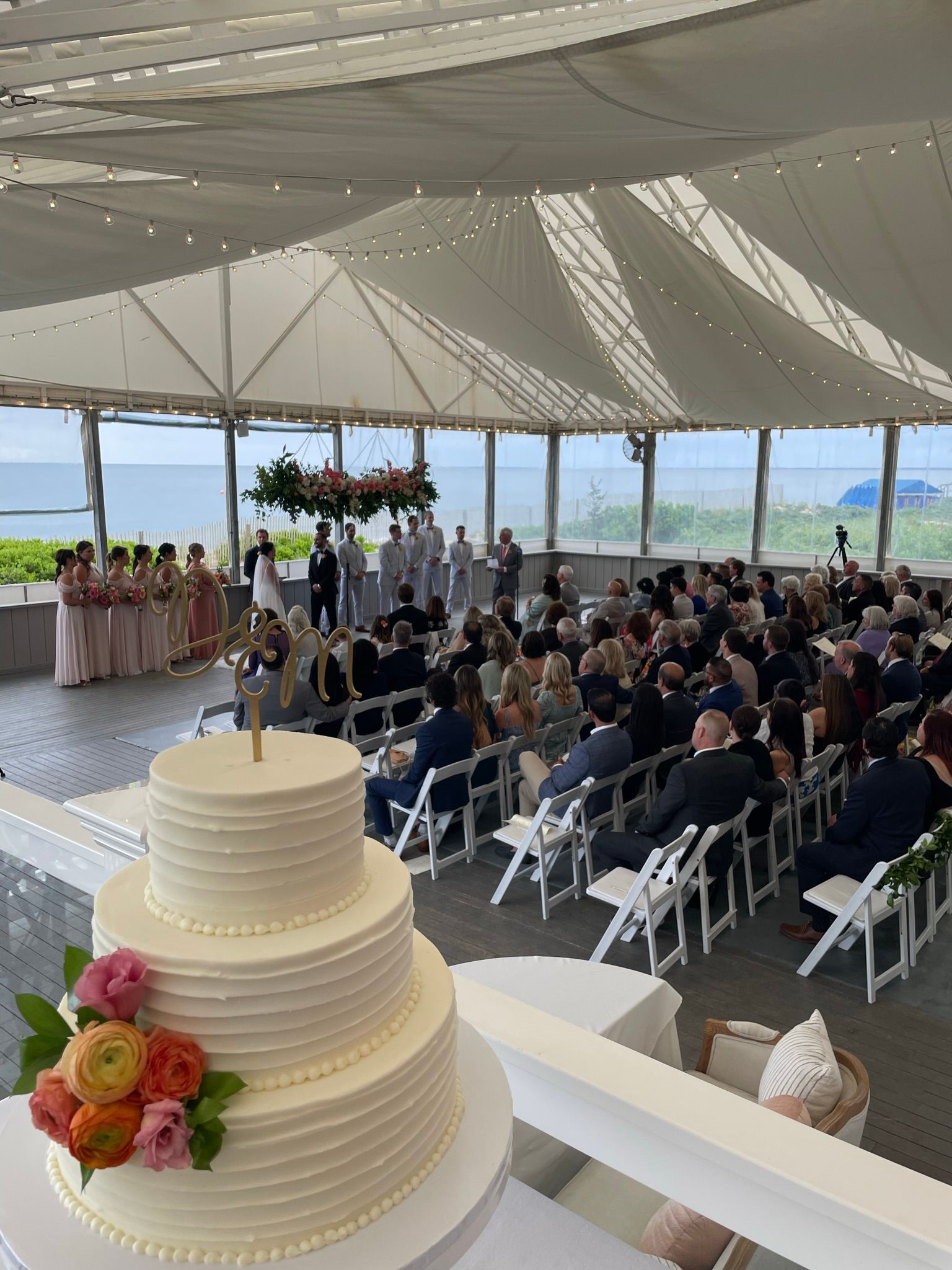 Wedding ceremony with a three-tier cake in the foreground and guests seated under a tent overlooking the water.