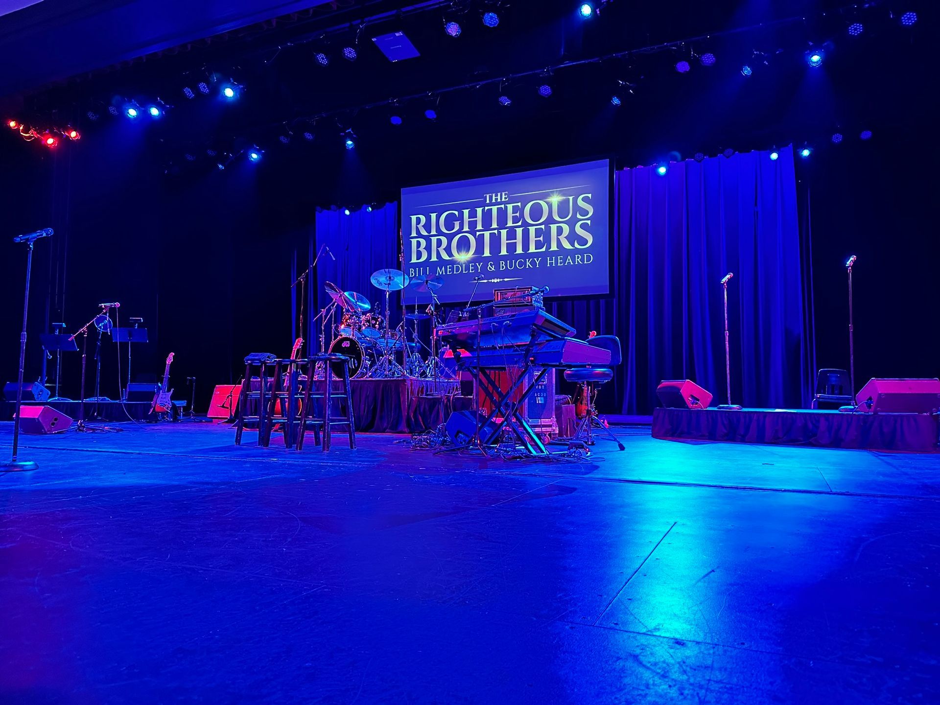 Stage setup for The Righteous Brothers concert with instruments, microphones, and a blue-lit background.