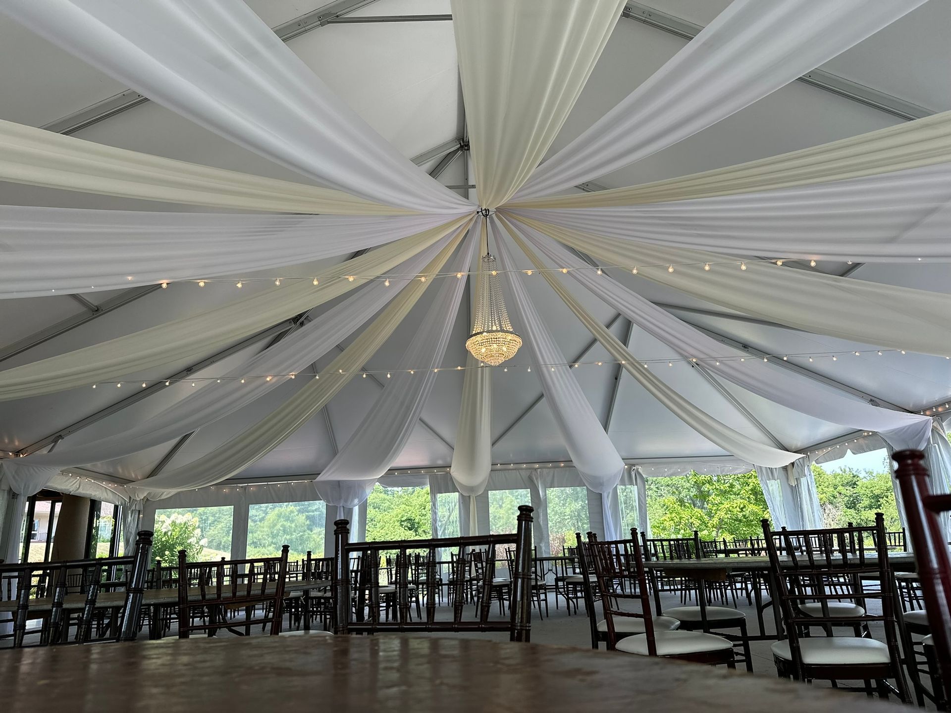 Indoor tent with white draped fabric ceiling, chandelier, tables, chairs, and outdoor views.