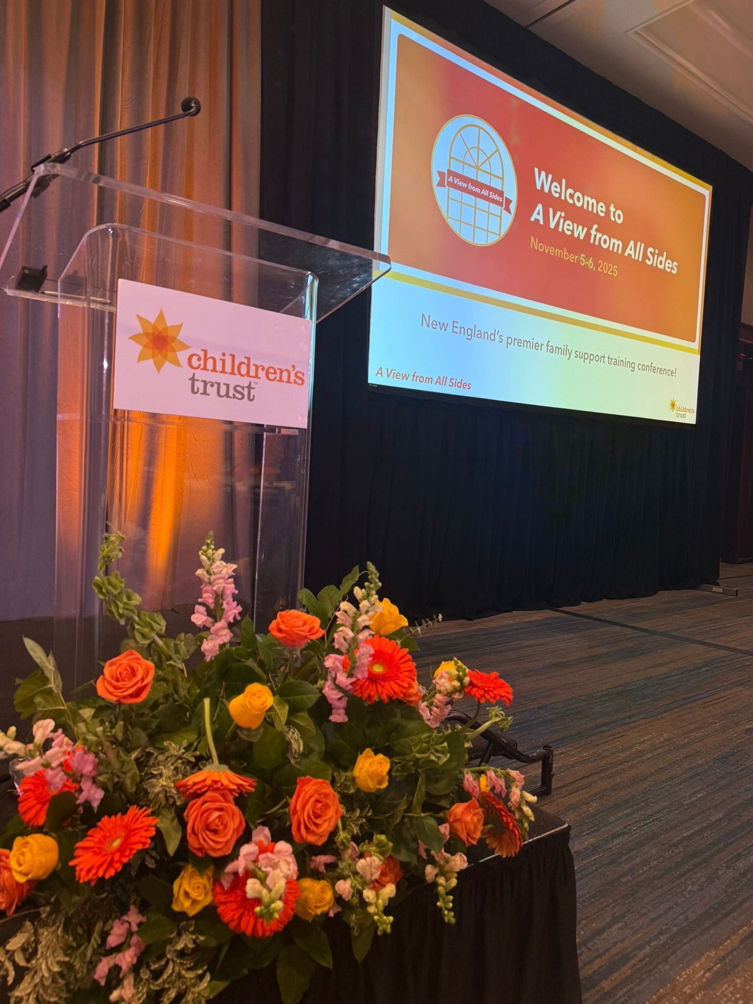 Podium with Children's Trust logo, floral arrangement, and a welcome slide at a conference.
