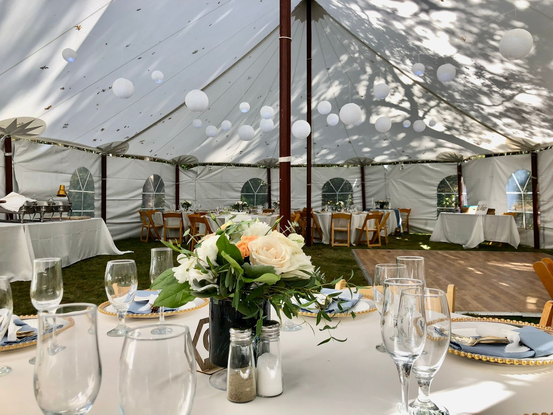 Wedding reception tent, tables set with flowers, dishes, and glassware; white paper lanterns hang from the ceiling.