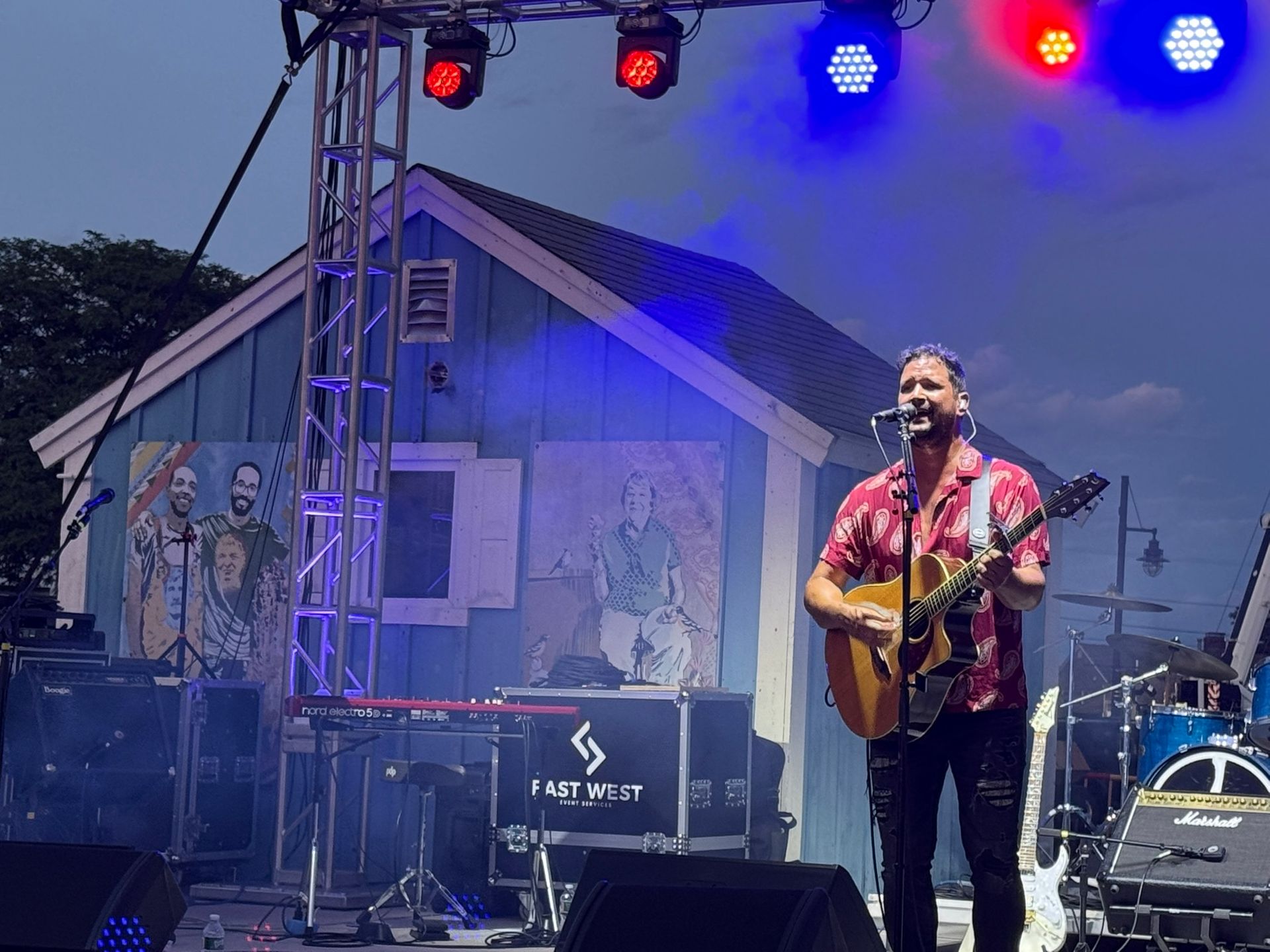 A man sings and plays guitar on stage, blue building backdrop, stage lights.