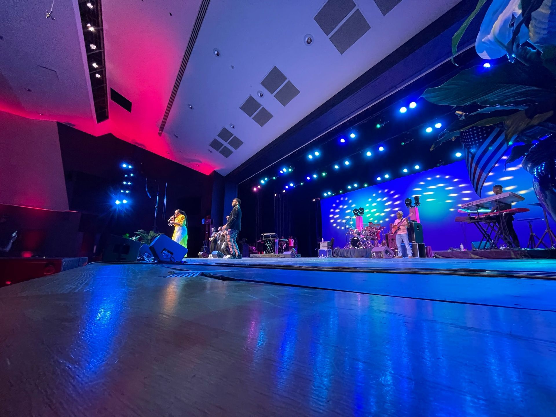 Band performing on a stage lit with blue and red lights. Musicians play instruments in a large auditorium.
