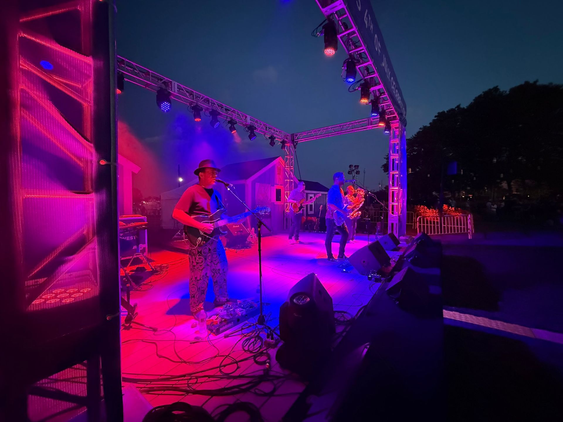 Band performing on a stage lit with blue and pink lights at dusk.