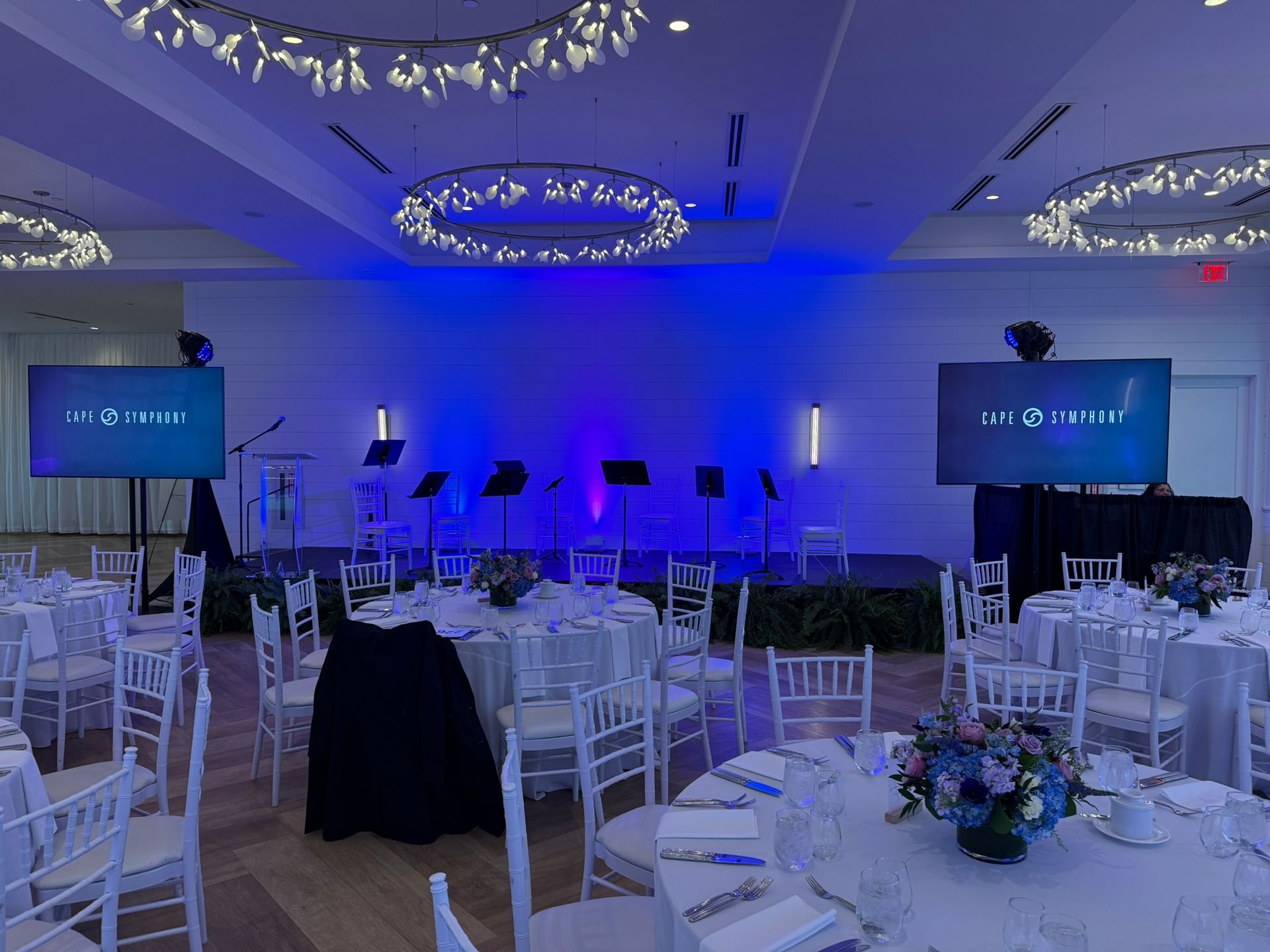 Event space with stage and tables. Blue lighting on the stage, tables set with white linens and chairs.