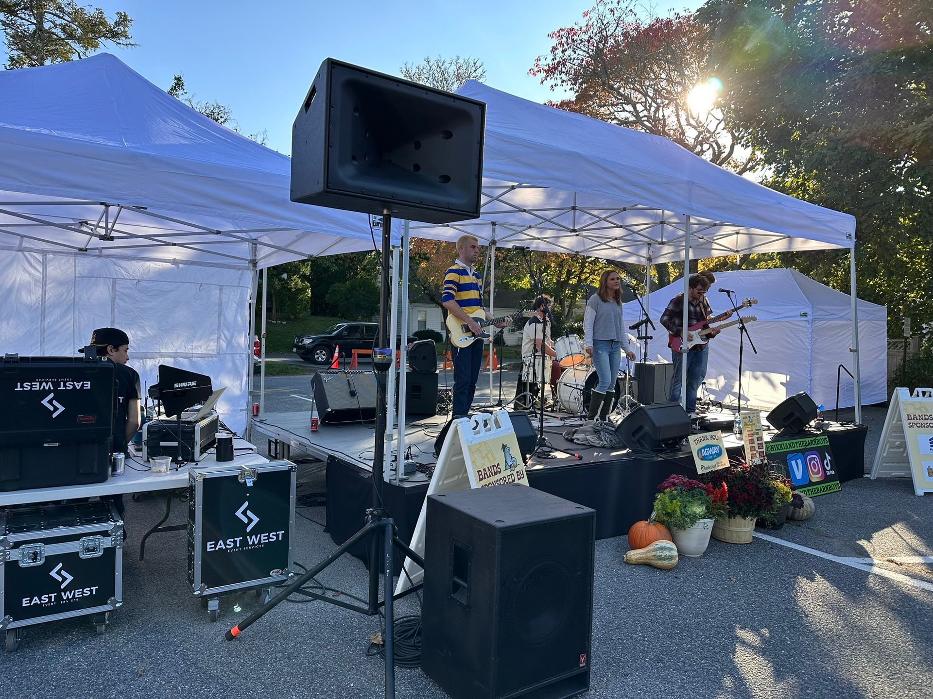 Outdoor band playing under tents at an event. Stage, sound equipment, and attendees are visible.