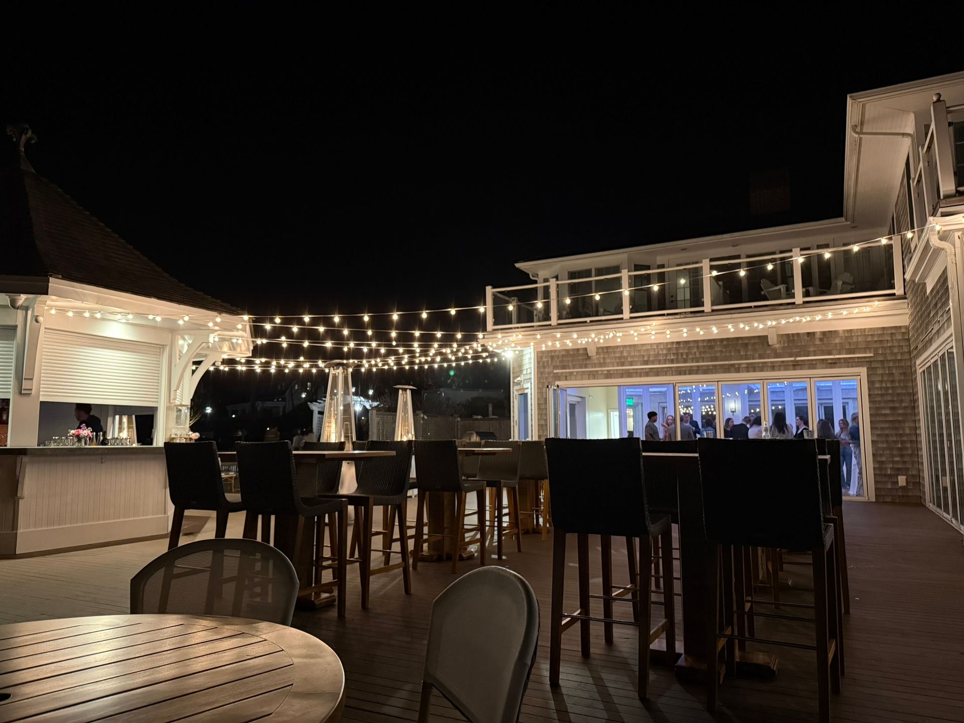 Outdoor patio bar at night with string lights, tall chairs, and building with open doors.
