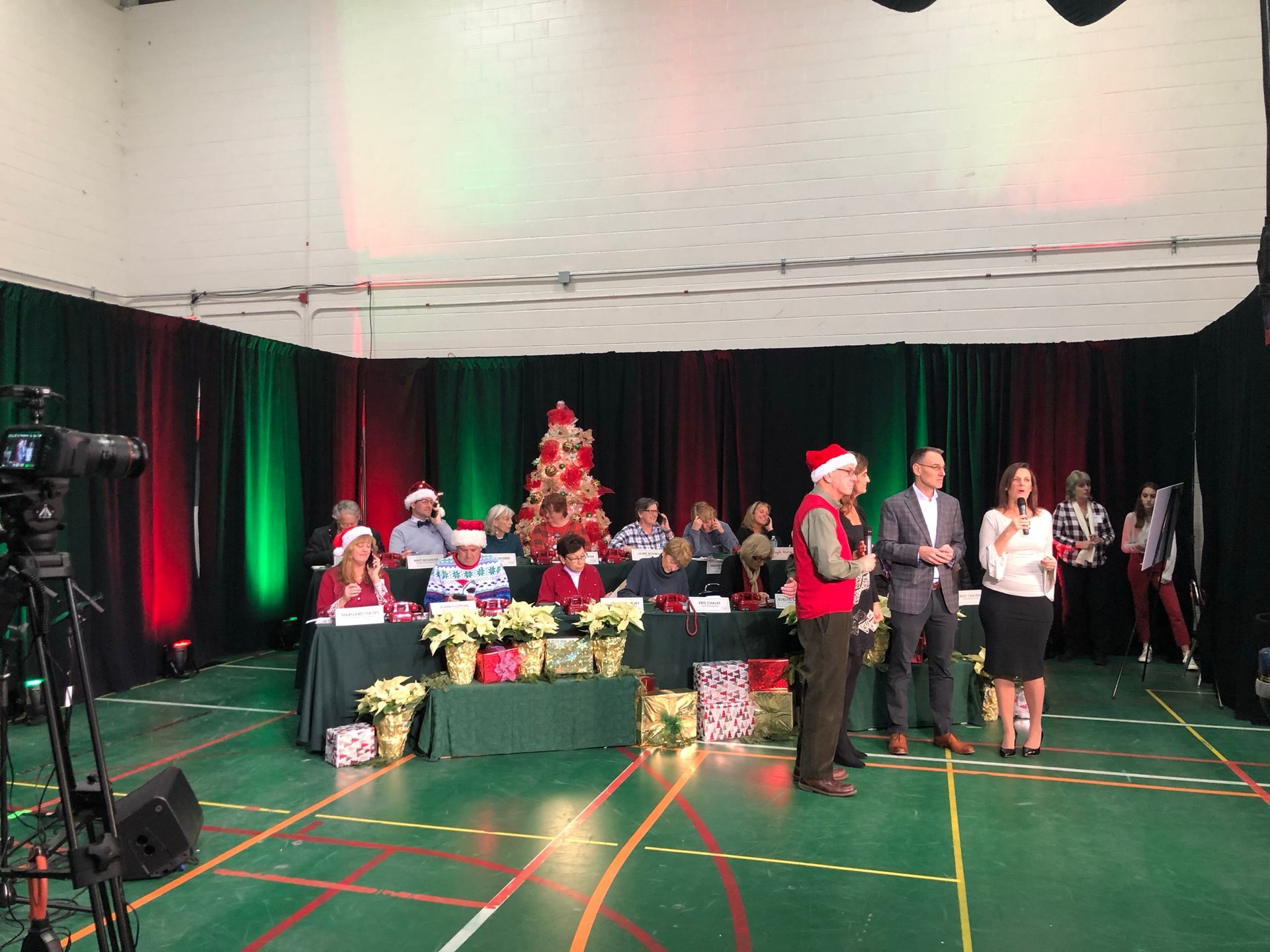 Christmas event: People on stage in front of holiday decorations. Some wearing Santa hats. Gifts and Christmas tree visible.