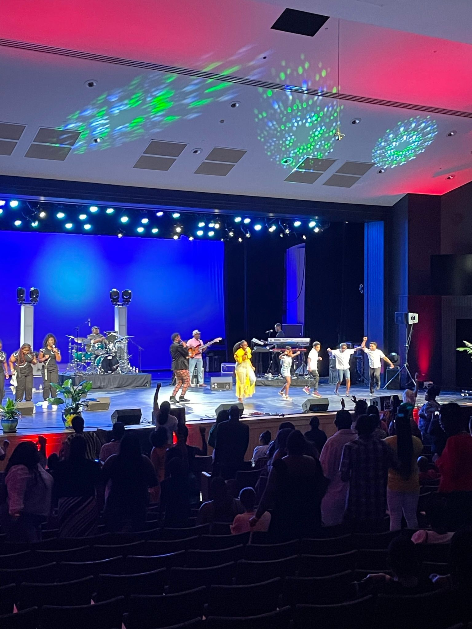Live music performance on a stage with a band, singers, and audience. Blue and red lighting.