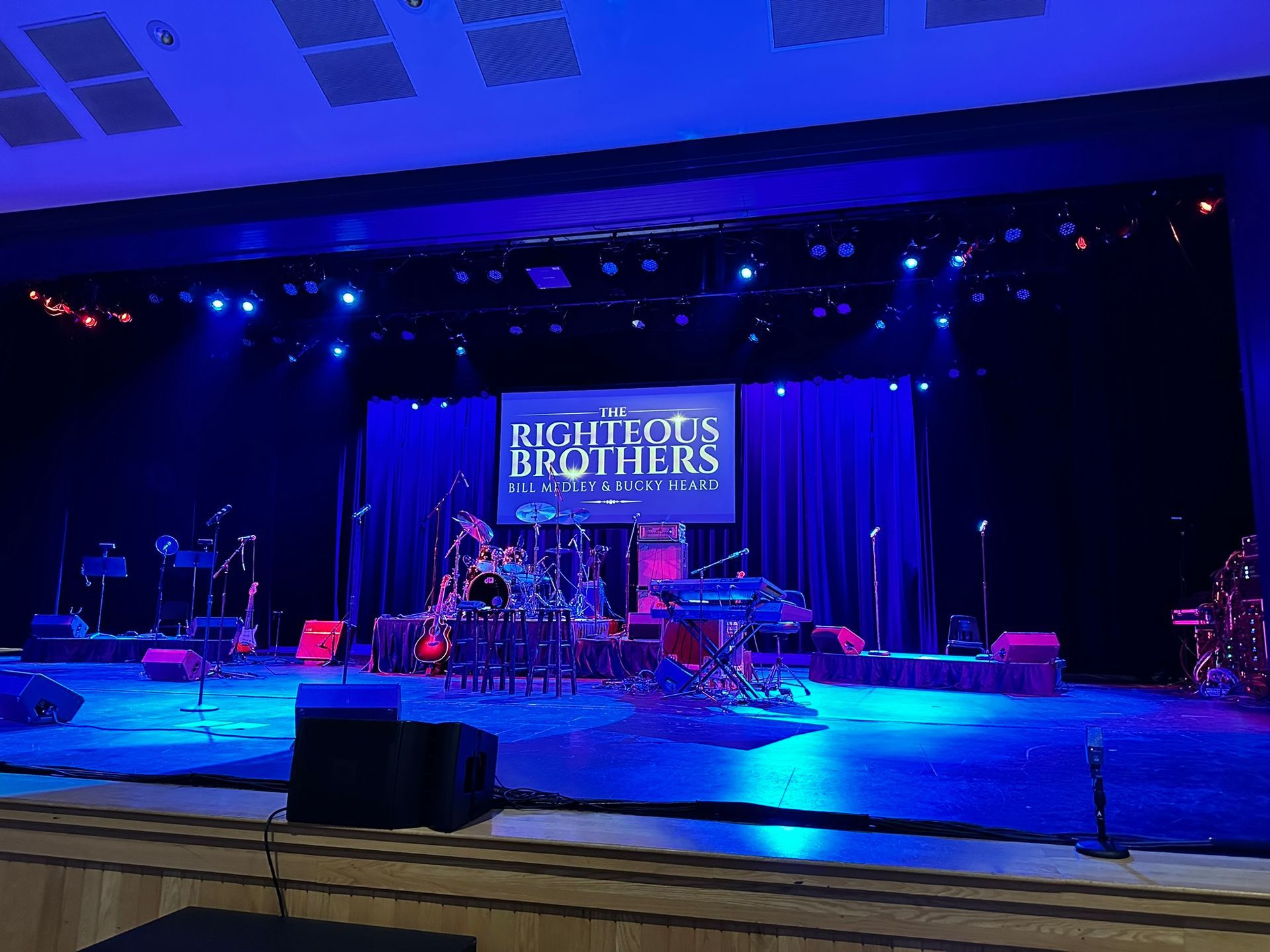 Stage set for a concert with blue lighting, drums, microphones, and a banner for the 