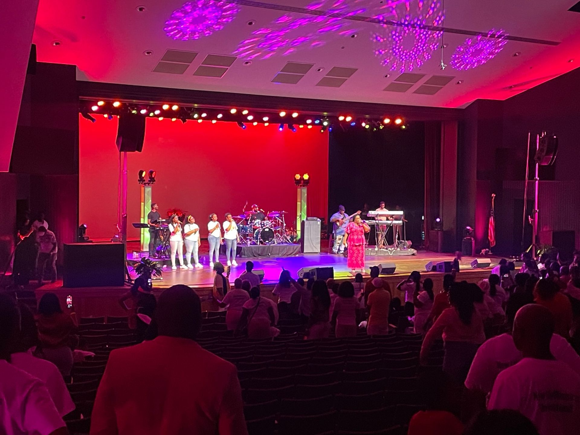 Concert stage with performers and audience in an auditorium lit with red and purple lights.