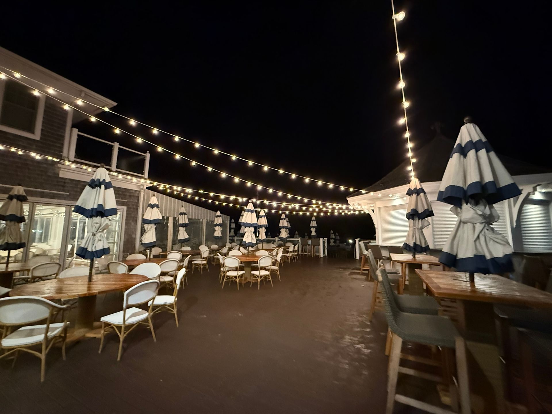 Outdoor restaurant patio at night, with string lights and blue-and-white umbrellas over tables and chairs.