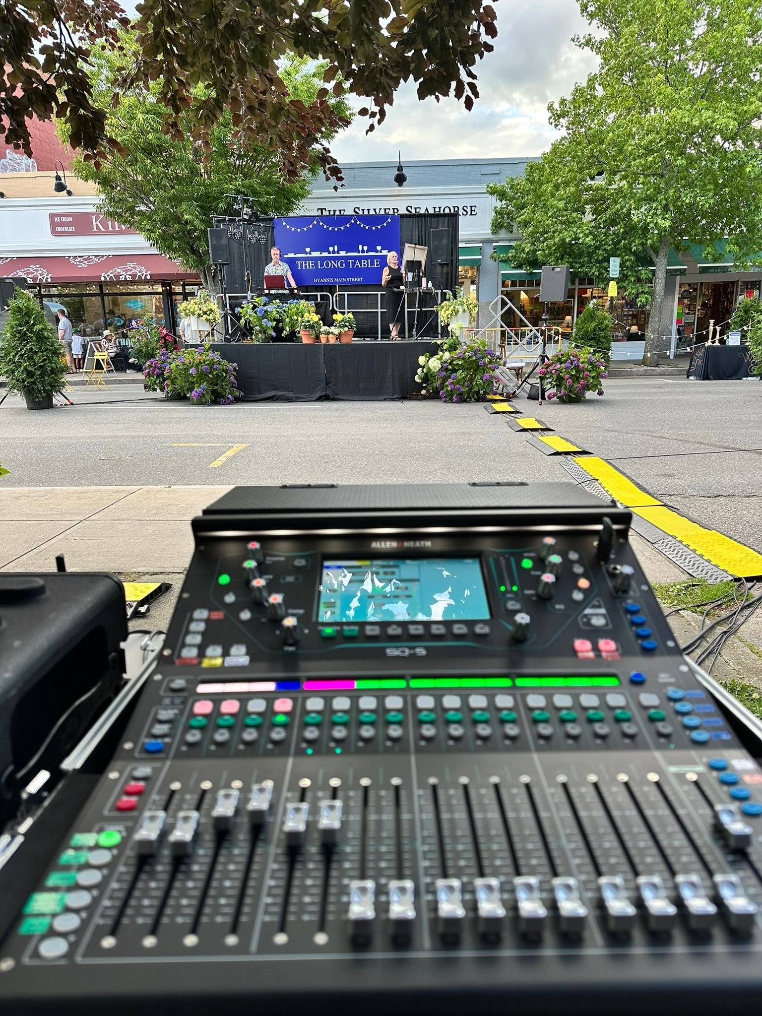 Soundboard in foreground with stage set up outdoors. Trees, buildings, and lake visible in the background.