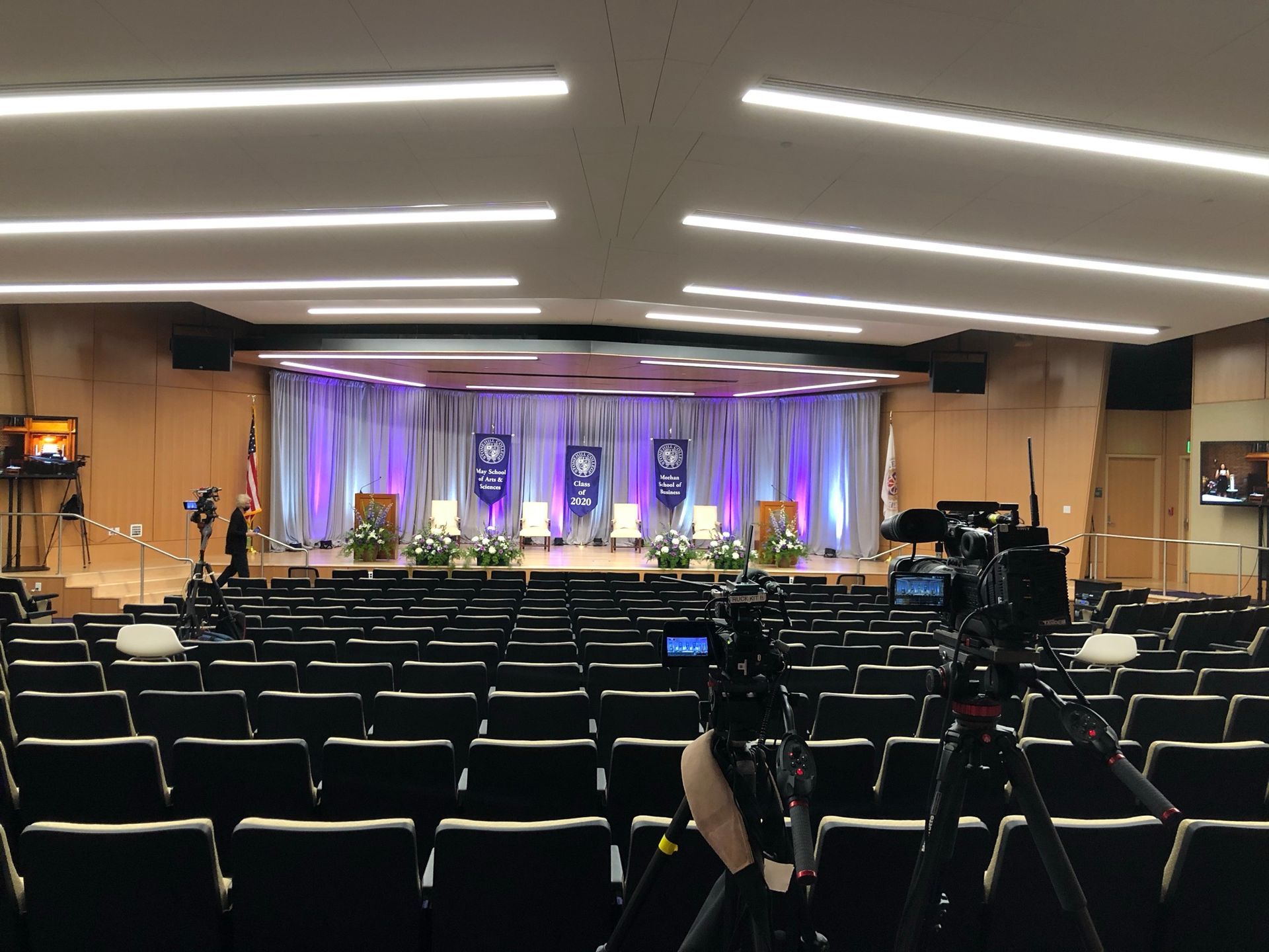 An auditorium stage set for a formal event with seating. Cameras are set up in the audience.