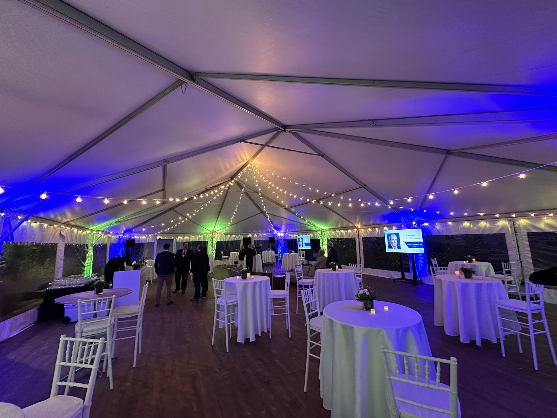 A tented event space with tables, chairs, and string lights, illuminated with blue and green lighting. People are mingling.