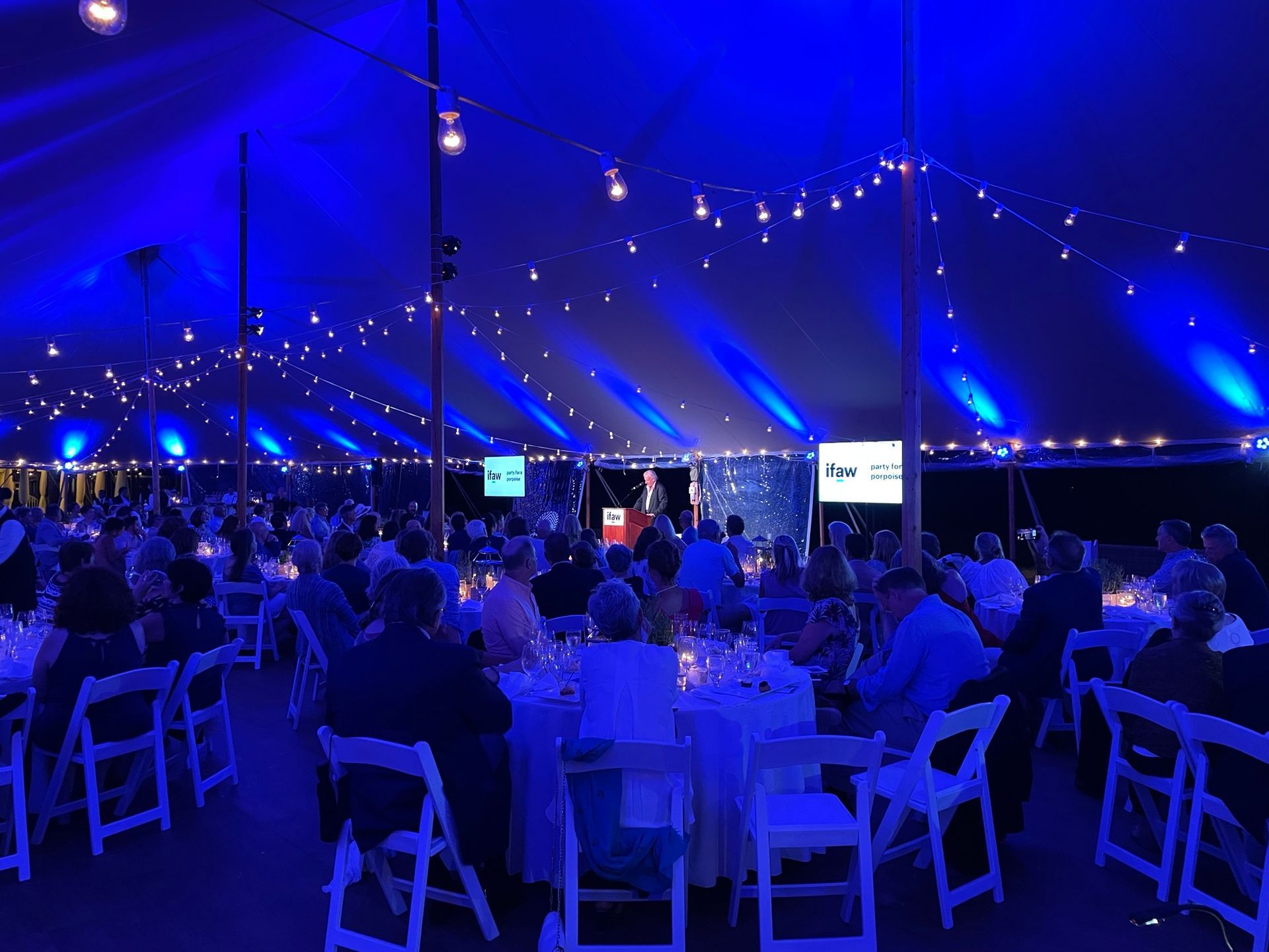 Event tent with blue lighting, people seated at tables, speaker at podium.