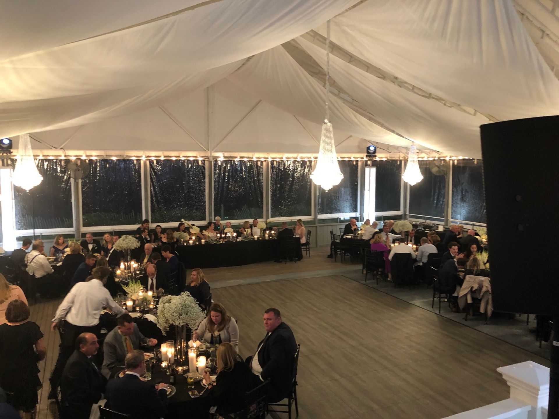Reception under a tent with guests seated at black-covered tables, chandeliers, and a wooden floor.