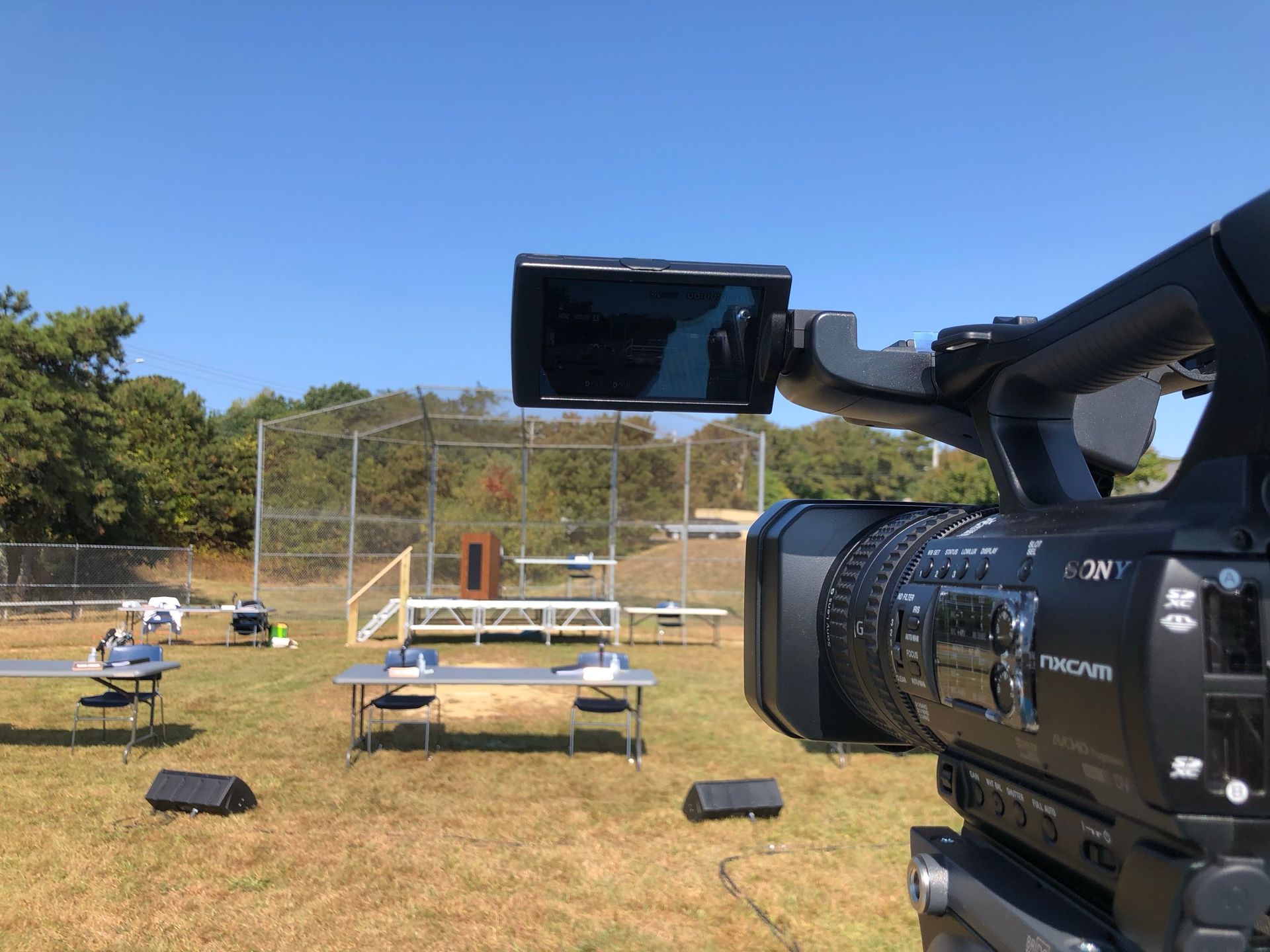Video camera setup outdoors filming a scene with a baseball backstop and tables.
