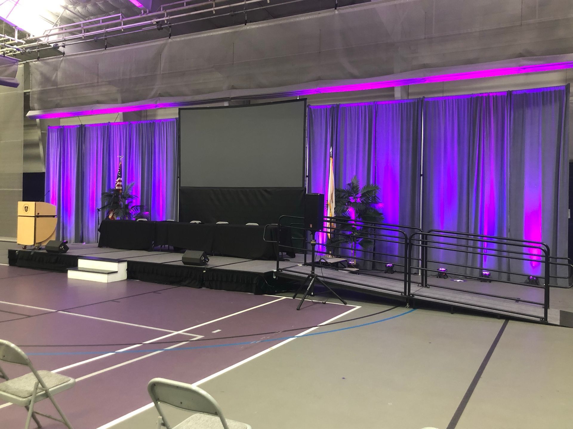 Stage with purple-lit curtains, screen, and plants, set up on a gymnasium floor.