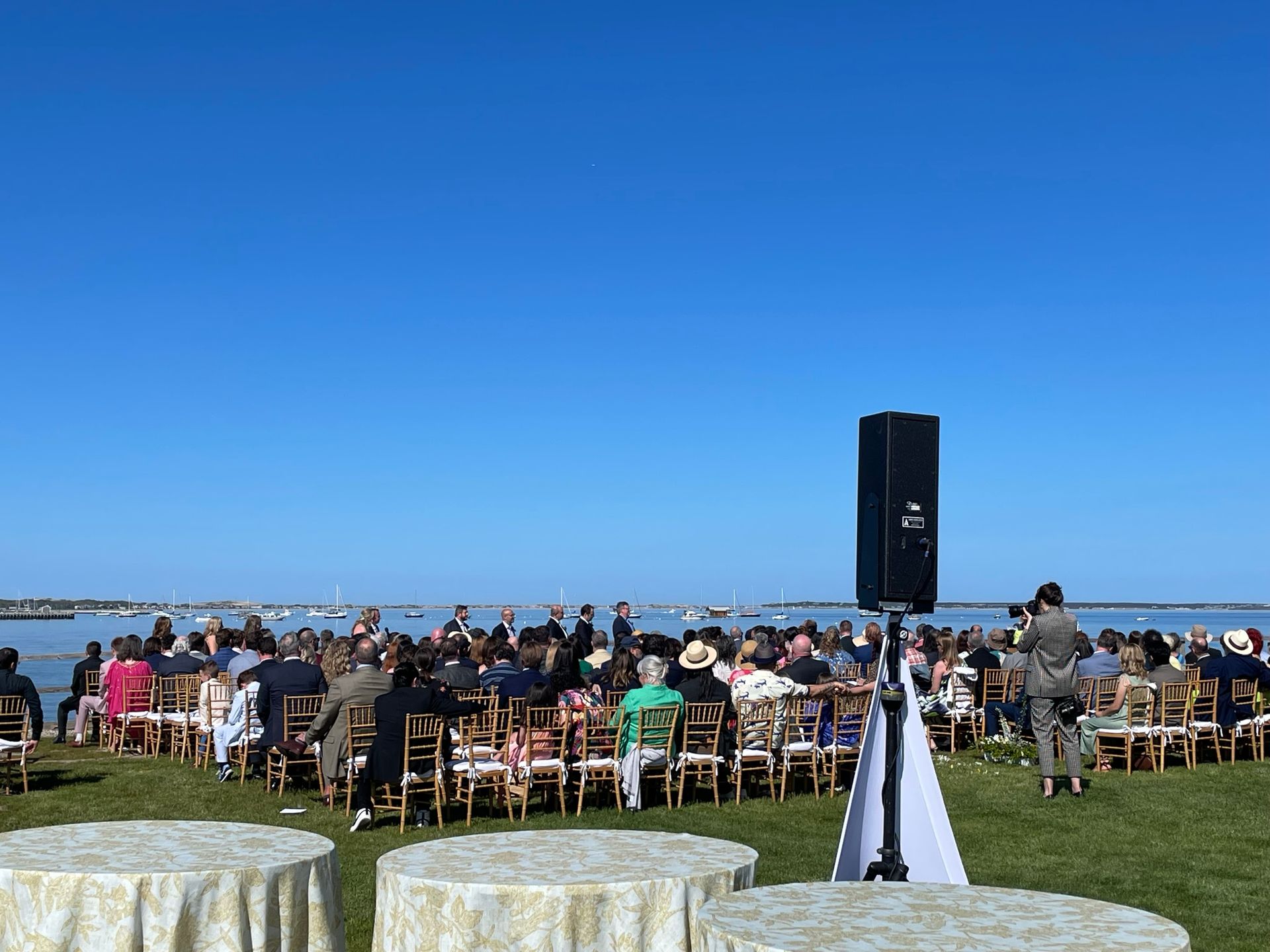 Outdoor wedding ceremony overlooking water. Guests seated, sound equipment set up, blue sky.