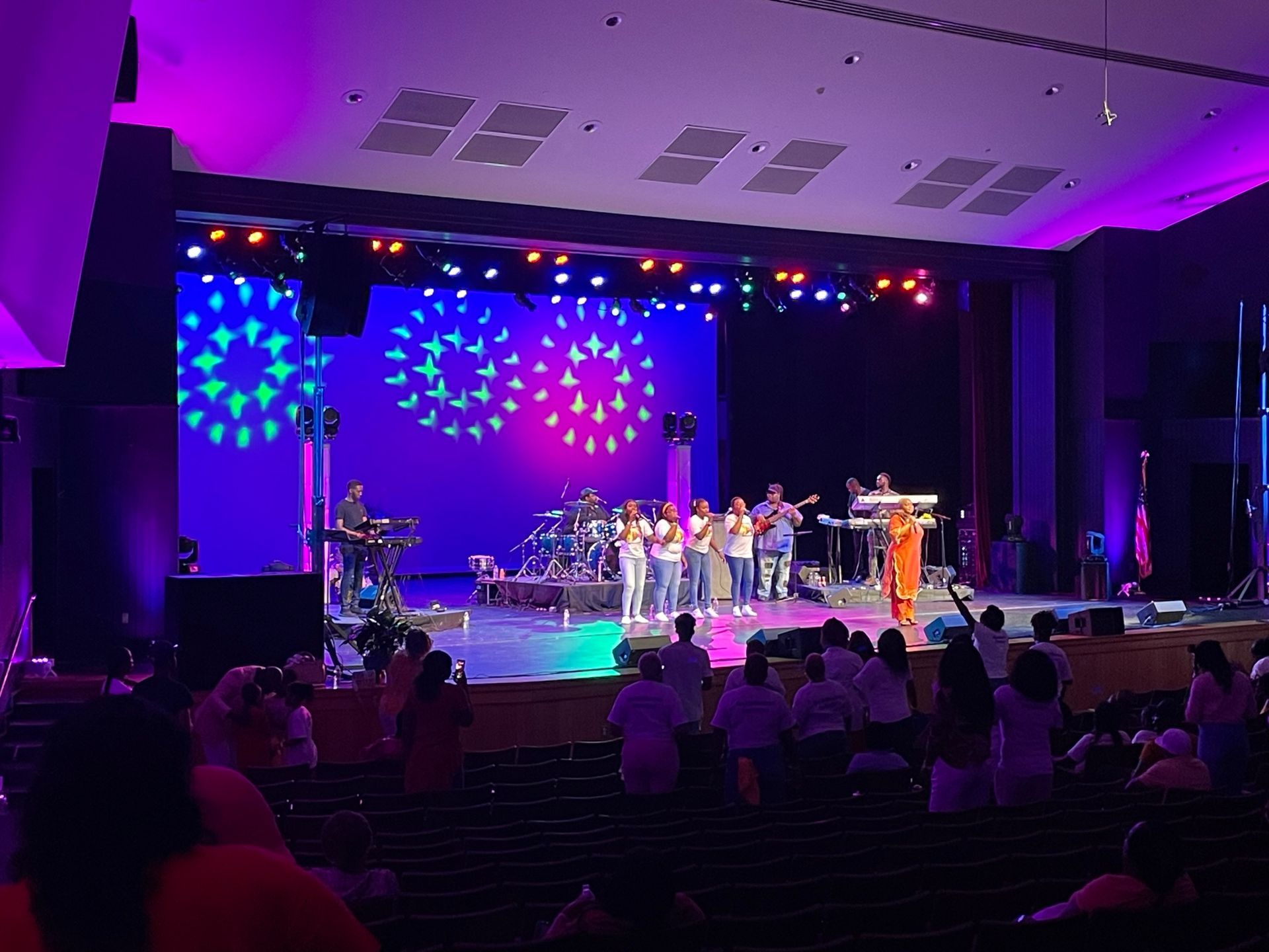 Musical performance on a stage with colorful lights. Audience seated in front.